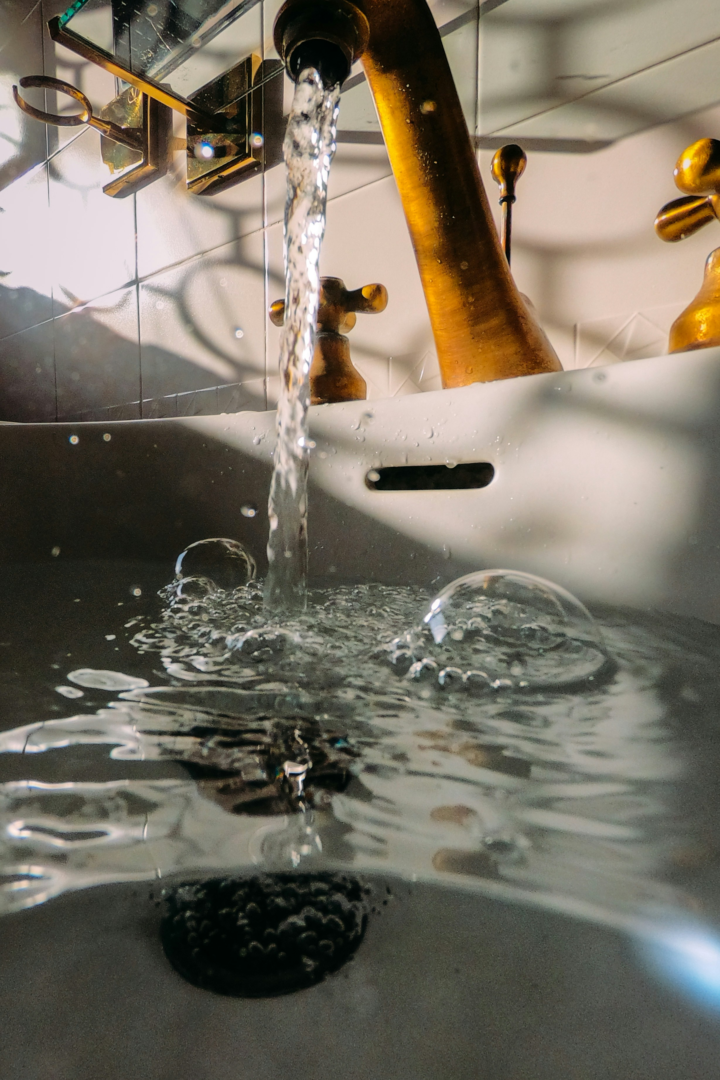 Close-up view of water flowing from a faucet into a sink, with a black scrub sponge at the bottom of the sink. The background shows a tiled wall with a mirror reflecting part of the faucet.