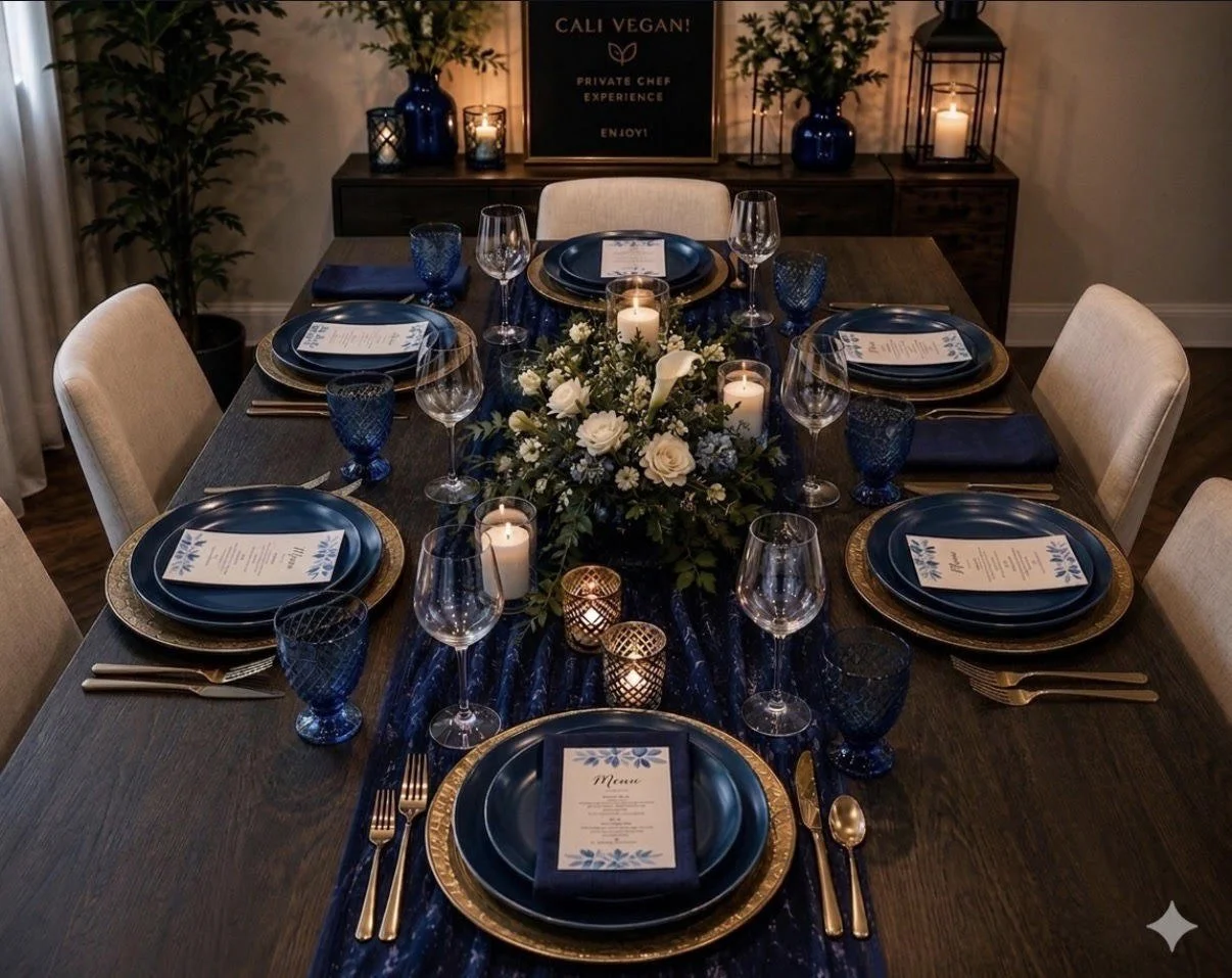 A elegantly set dining table with six place settings, featuring blue plates with gold rims, gold utensils, blue wine glasses, and printed menus. The center of the table has a green foliage arrangement with white flowers and candles, creating a sophisticated ambiance. In the background, there is a sign that reads 'CALI VEGAN! Private Chef Experience Enjoy' on a dark sideboard, with matching large blue vases, candles, and decorative lanterns.
