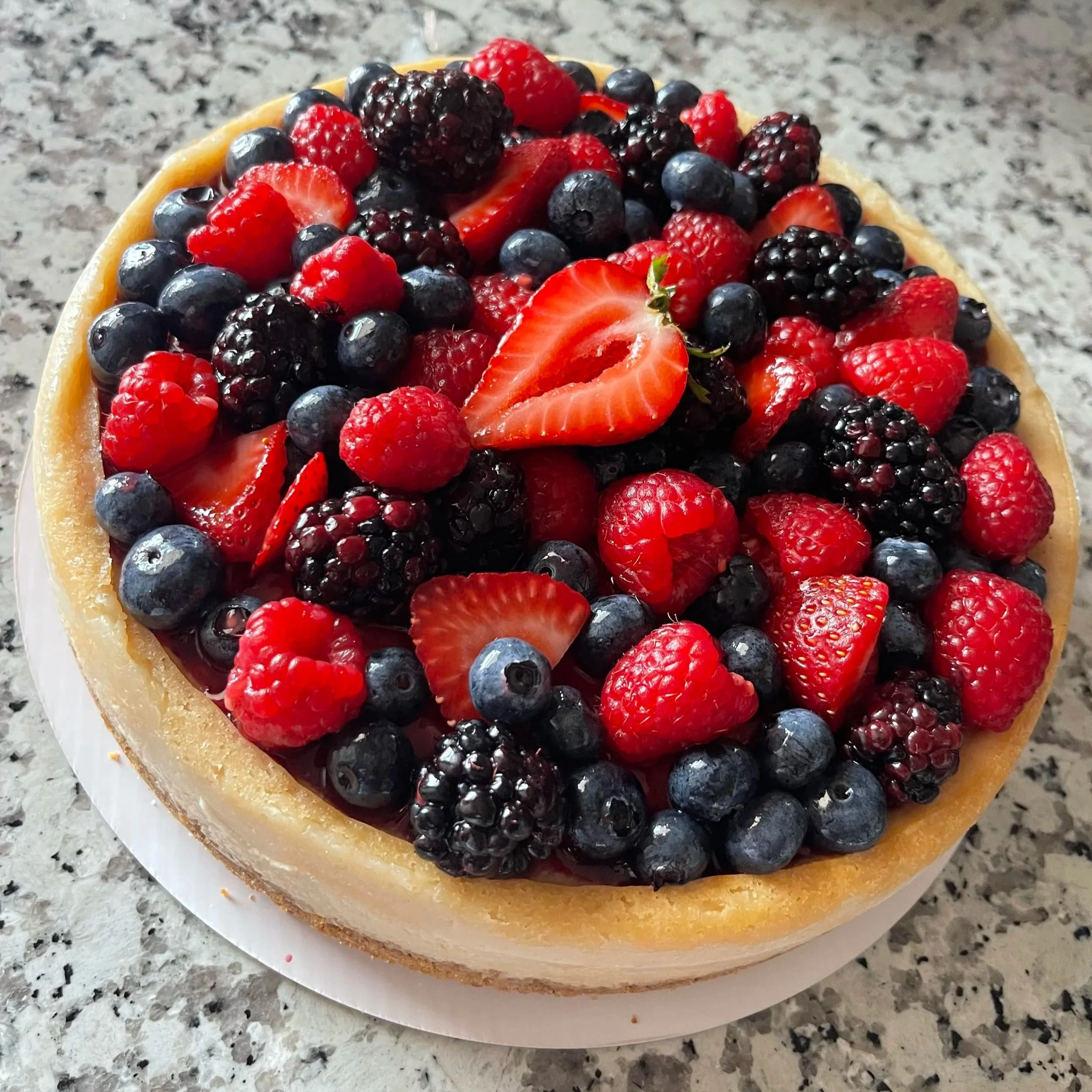 A very berry cheesecaske topped with blueberries, strawberries, Raspberries, and blackberries on a gray speckled countertop.