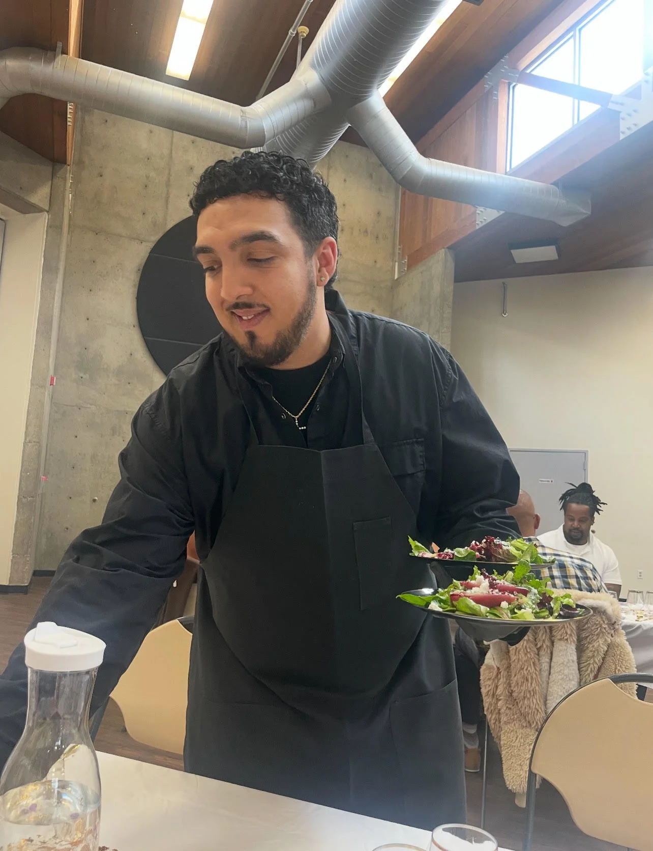 A young man with curly black hair and a beard, wearing a black apron, is serving a salad at a restaurant. The background includes an industrial-style ceiling with exposed ducts and large windows, along with other diners.