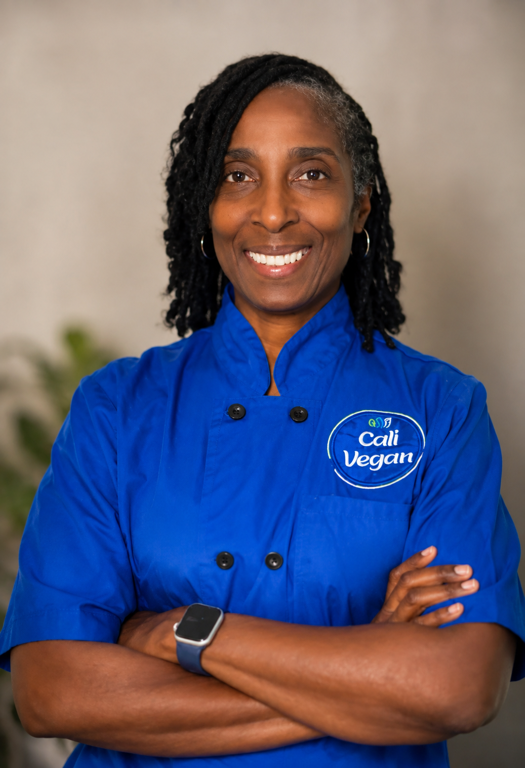 A woman wearing a blue chef's coat with a 'Cali Vegan' logo, smiling with arms crossed, in an indoor setting.