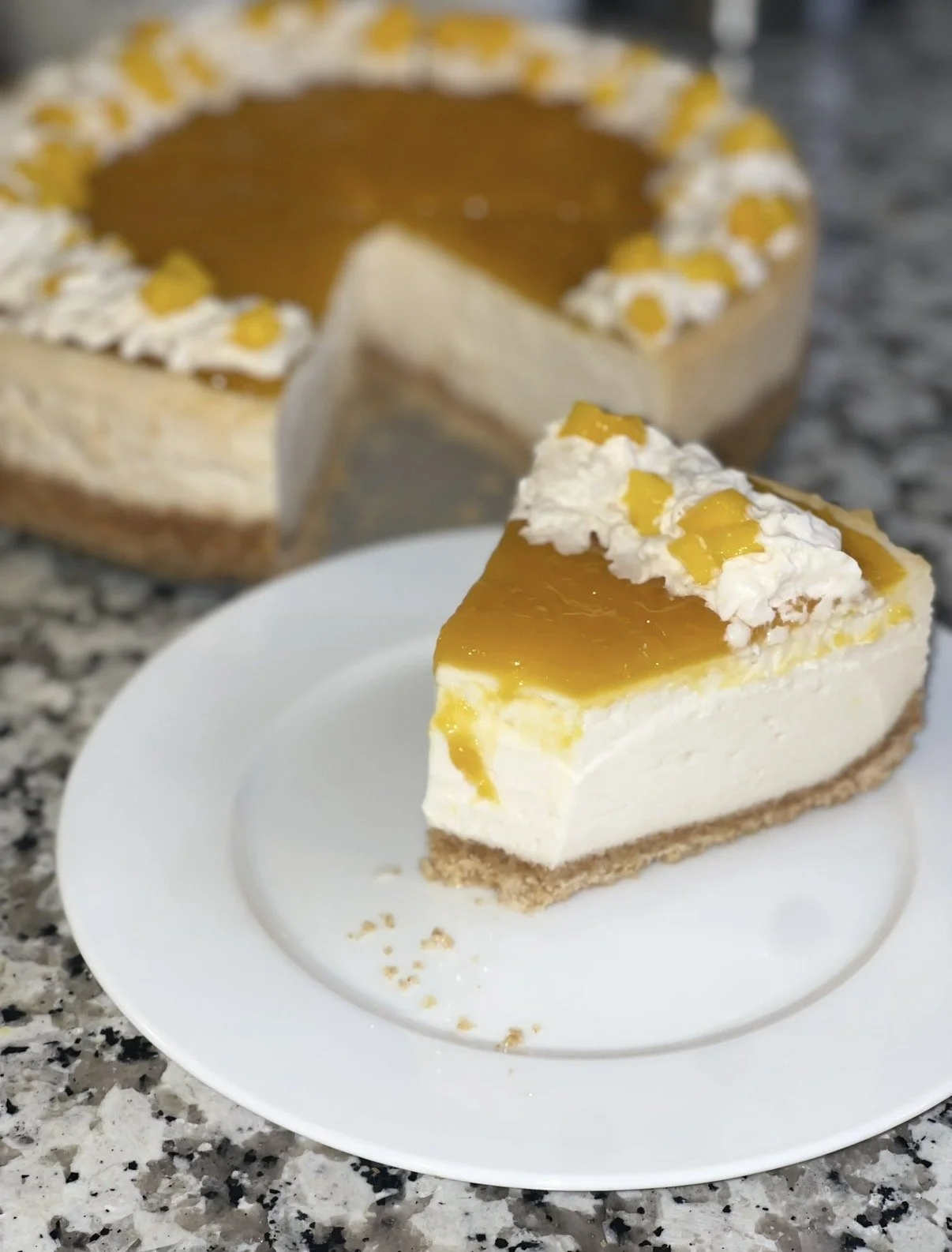 A slice of mango cheesecake on a white plate, with a graham cracker crust, and whipped cream, with the remaining cheesecake in the background.