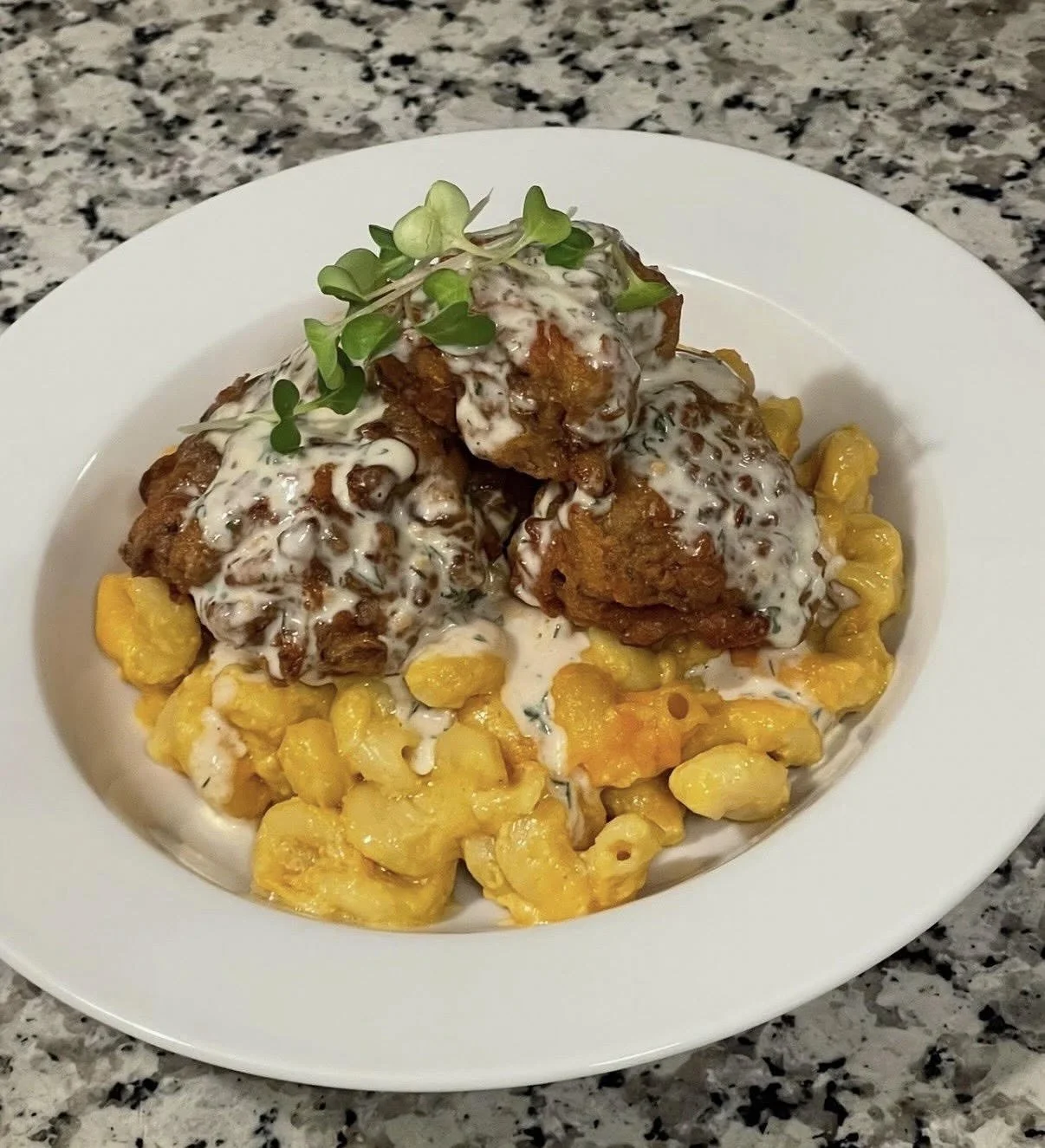 Fried chicken drumsticks topped with creamy white sauce and microgreens, served over macaroni and cheese on a white plate, with a granite countertop background.