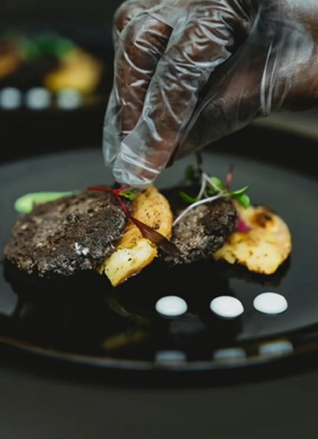 Close-up of a chef's hand in a glove garnishing a potato and mushroom patty Napoleon dish on a black plate with small white sauce dots and microgreens