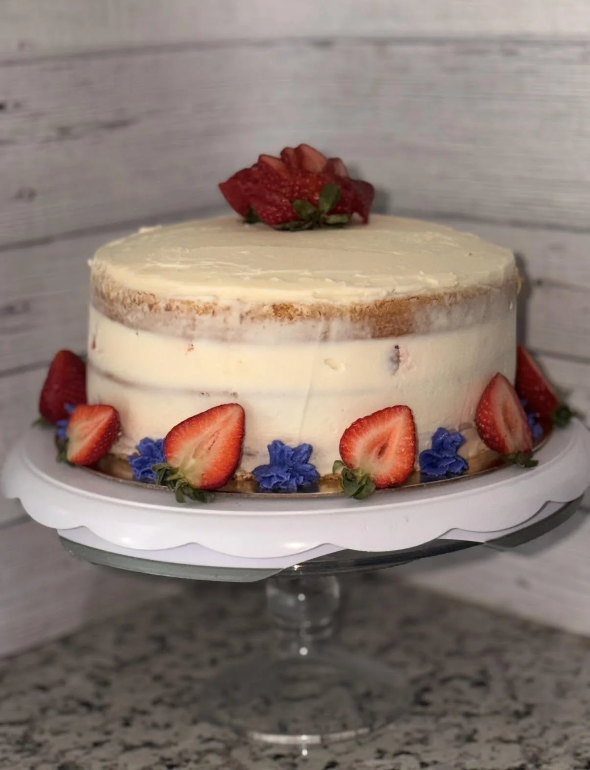 A layered cake with white frosting, decorated with sliced strawberries and small purple flowers around the base, topped with a halved strawberry on top.