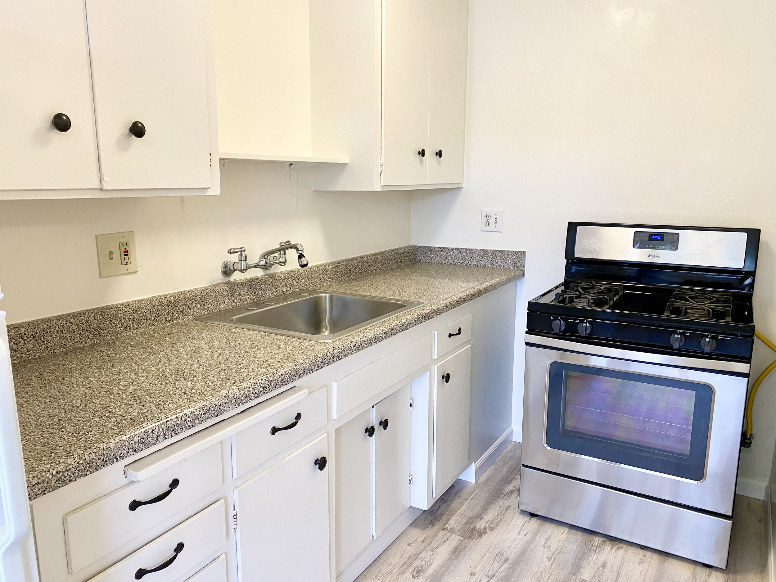 A kitchen with white cabinets, a speckled countertop, a stainless steel sink, and a Black and stainless steel stove with an oven. There are electrical outlets above the counter.