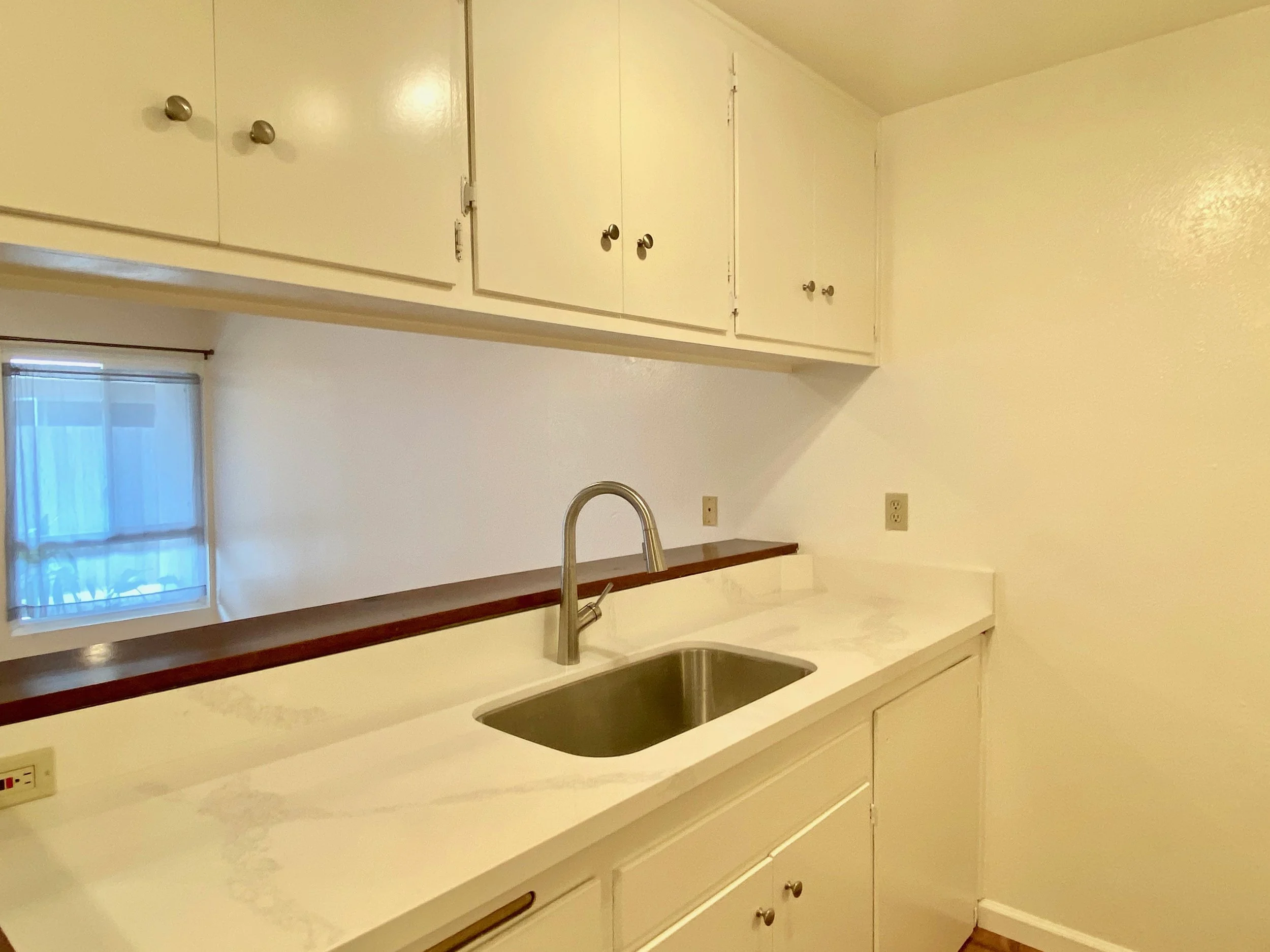 A white kitchen with a countertop, a stainless steel sink, a faucet, and upper cabinets with round knobs. A window with sheer curtains is visible in the background.