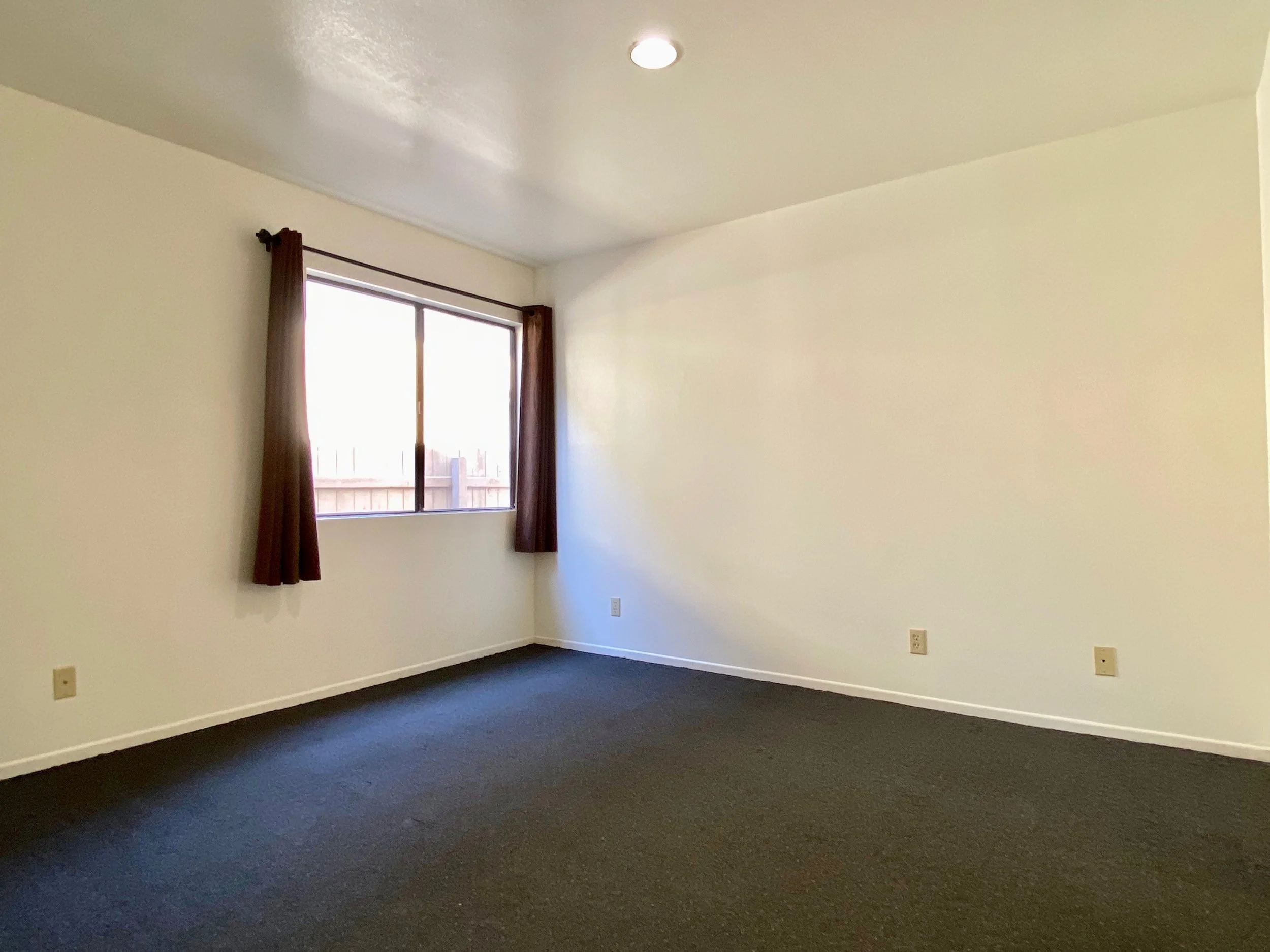 Empty room with beige walls, dark carpeting, a window with brown curtains, and visible electrical outlets.