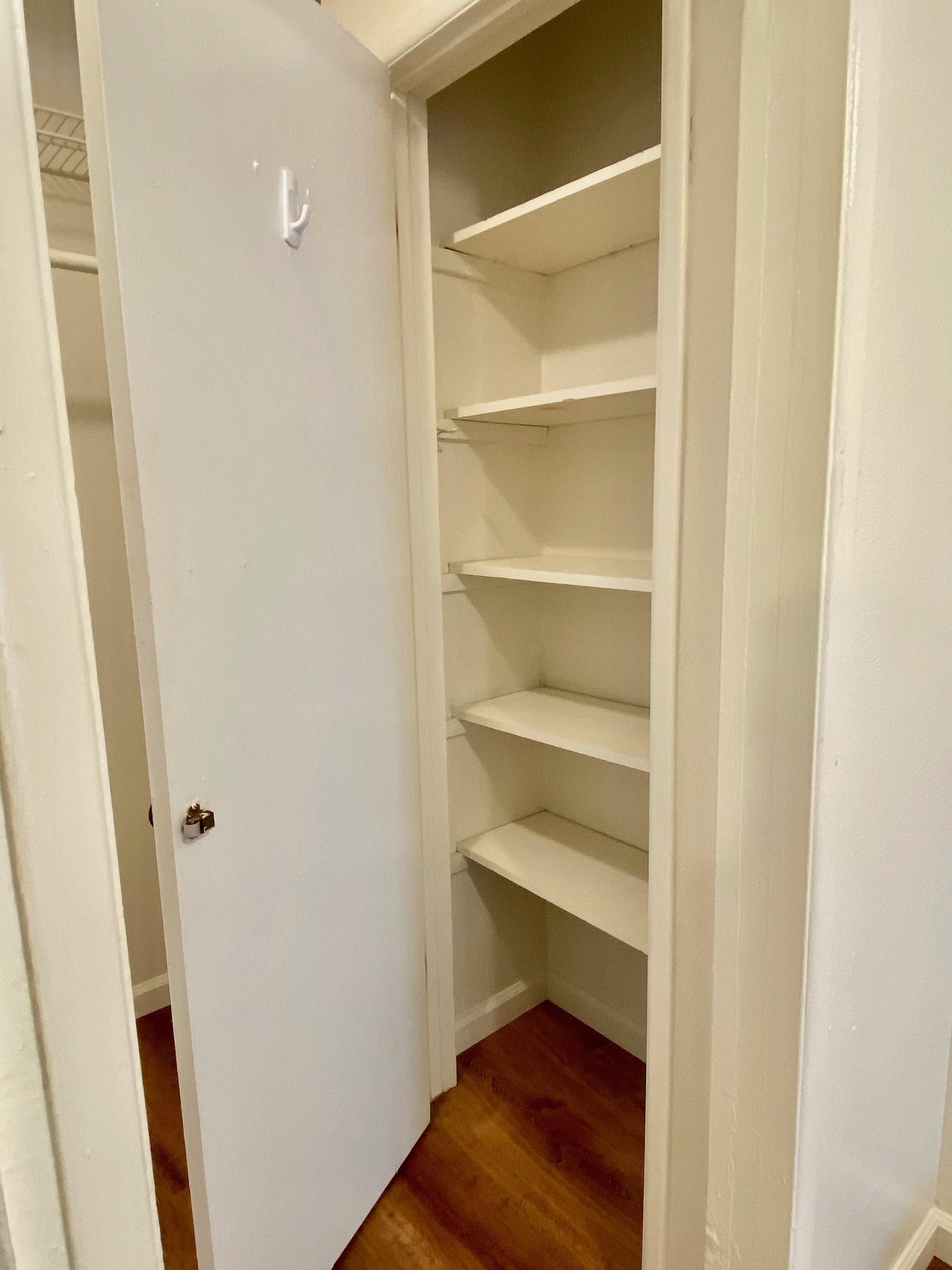 Empty built-in closet with white shelves and door, located on a wooden floor.