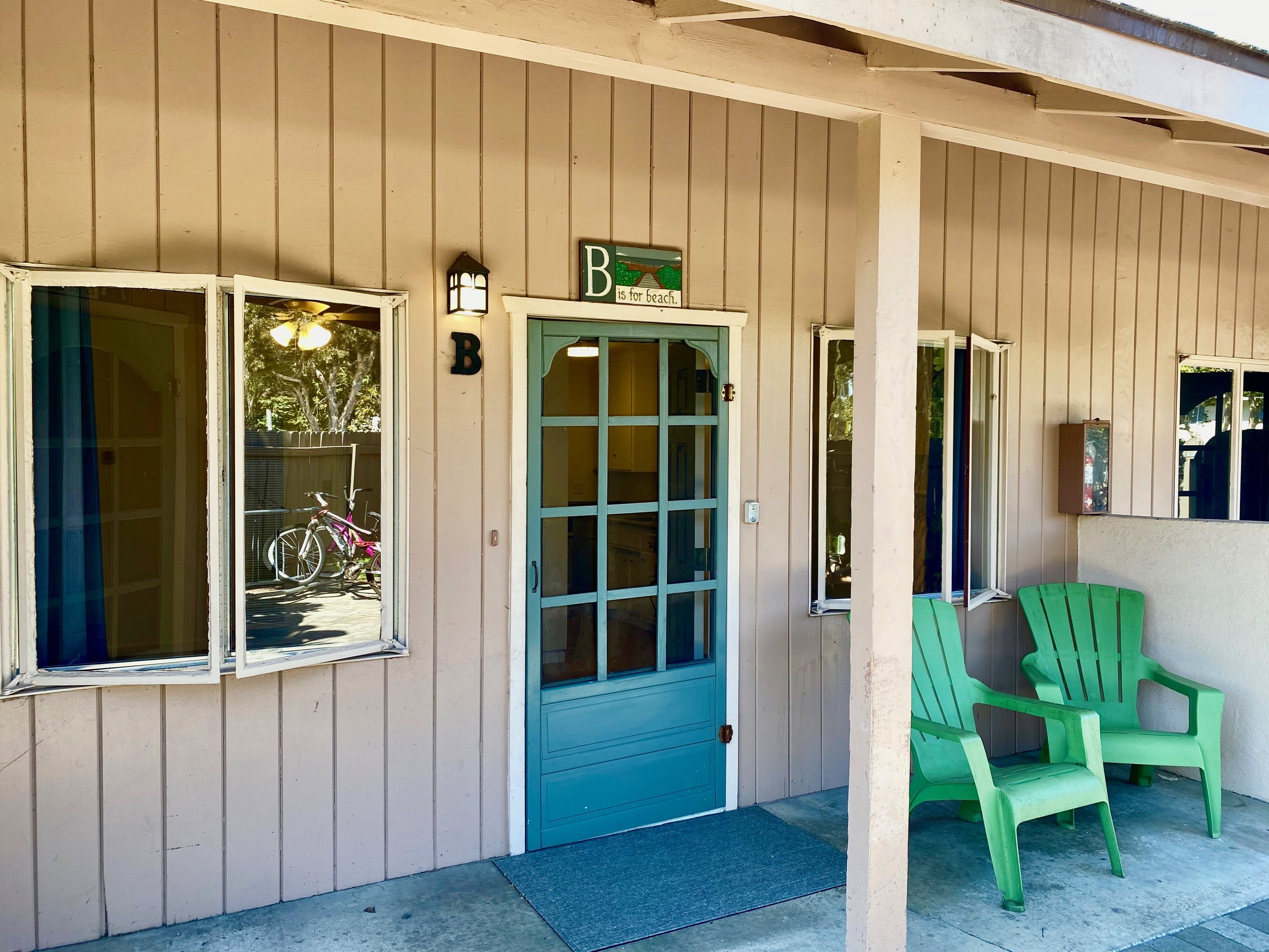 A close-up of a house exterior with beige vertical siding, a blue door, and an open window. There are two green chairs next to the wall and a pink bicycle reflected in the window. A sign above the door reads, 'B is for beach' with a beach scene illus
