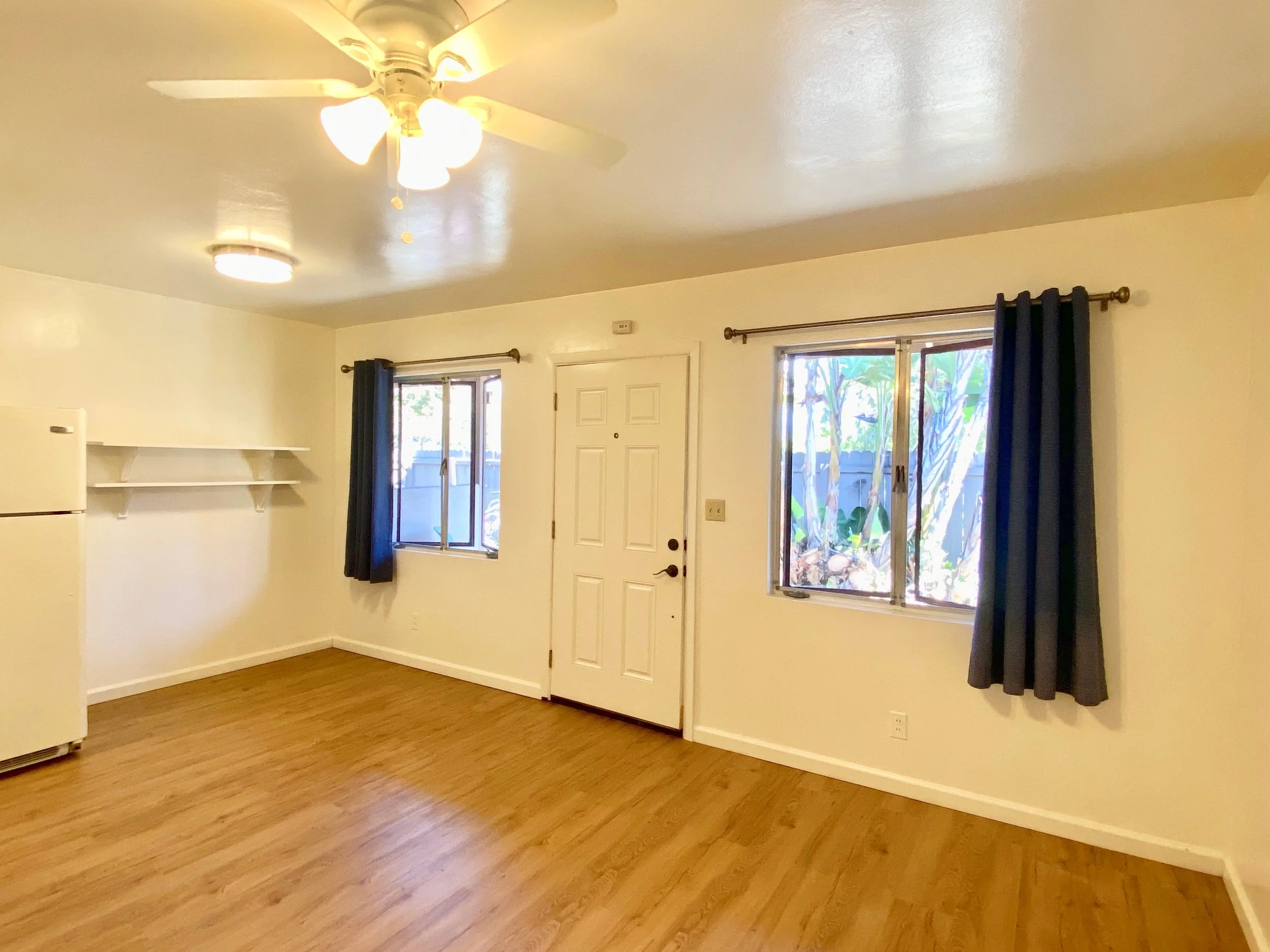 A bright living room with white walls, wood floor, a ceiling fan with lights, two windows with dark curtains, a white door, and a small white refrigerator.