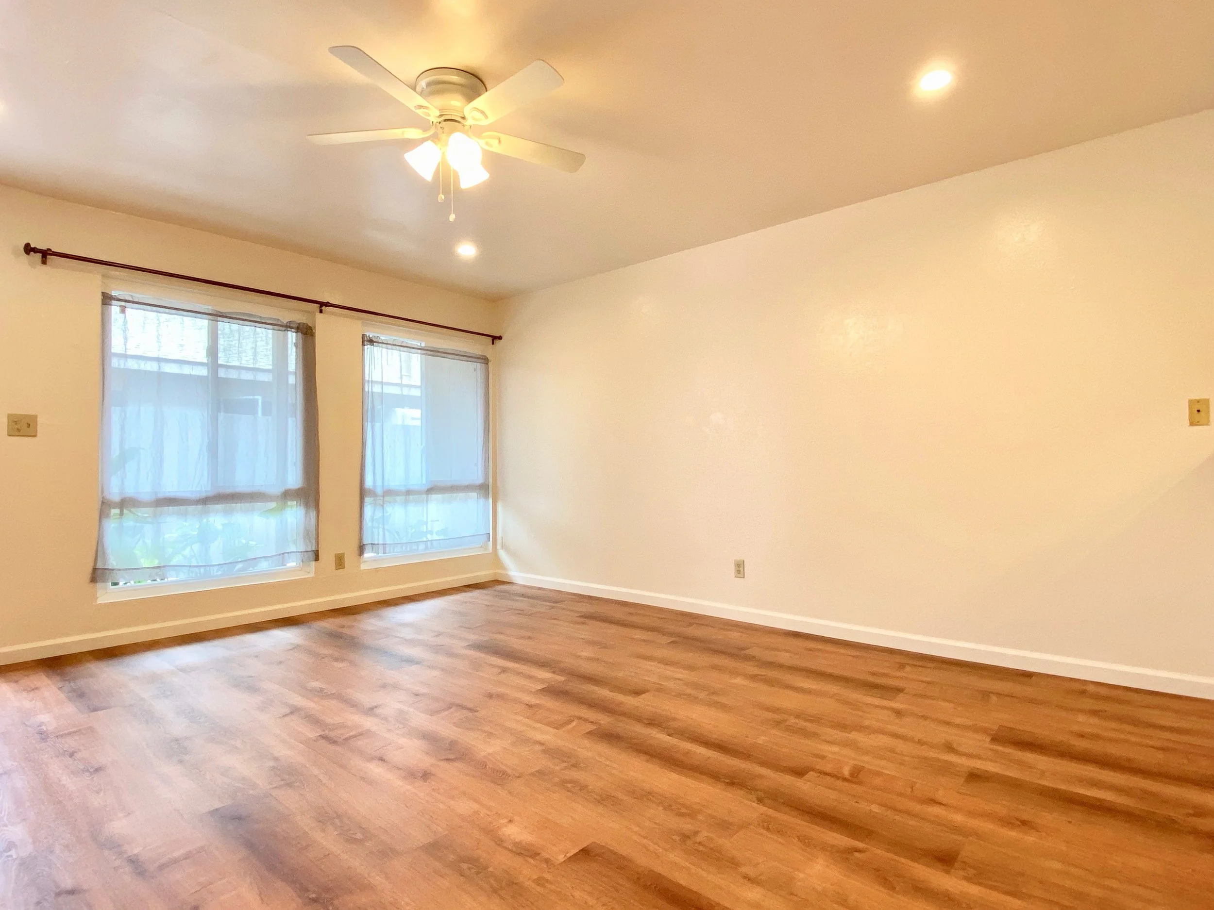 Empty room with hardwood flooring, white walls, two windows with sheer curtains, ceiling fan with lights, and recessed lighting.