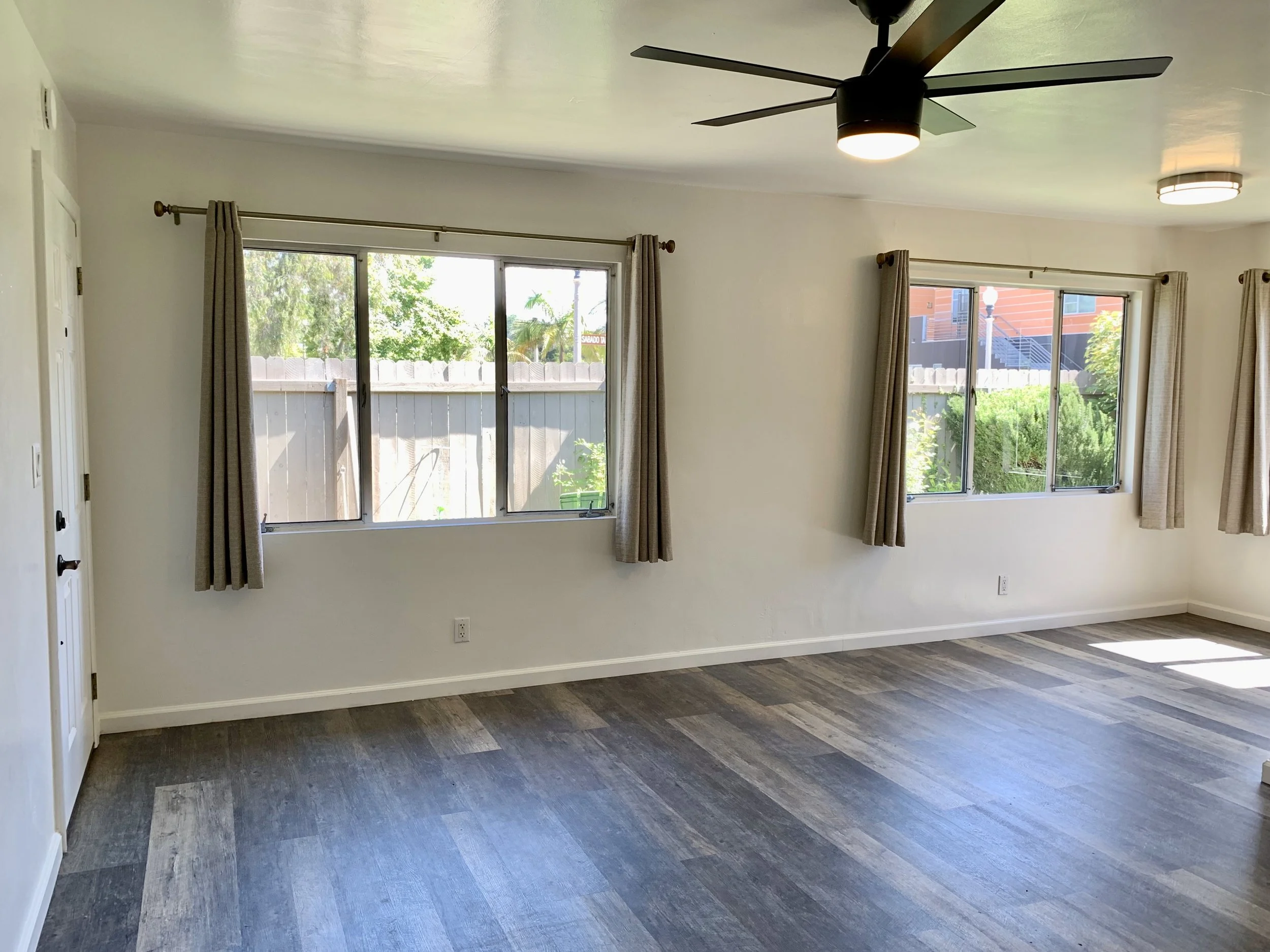 Empty living room with two large windows, beige curtains, wood flooring with a dark plank pattern, and a black ceiling fan with a light.