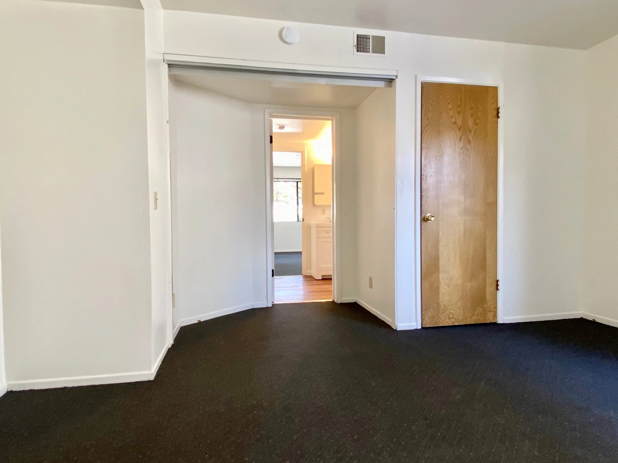 Interior of an empty room with dark carpet flooring, white walls, a wooden door on the right, and a doorway leading to a lit room with a window. The ceiling has a smoke detector and an air vent.