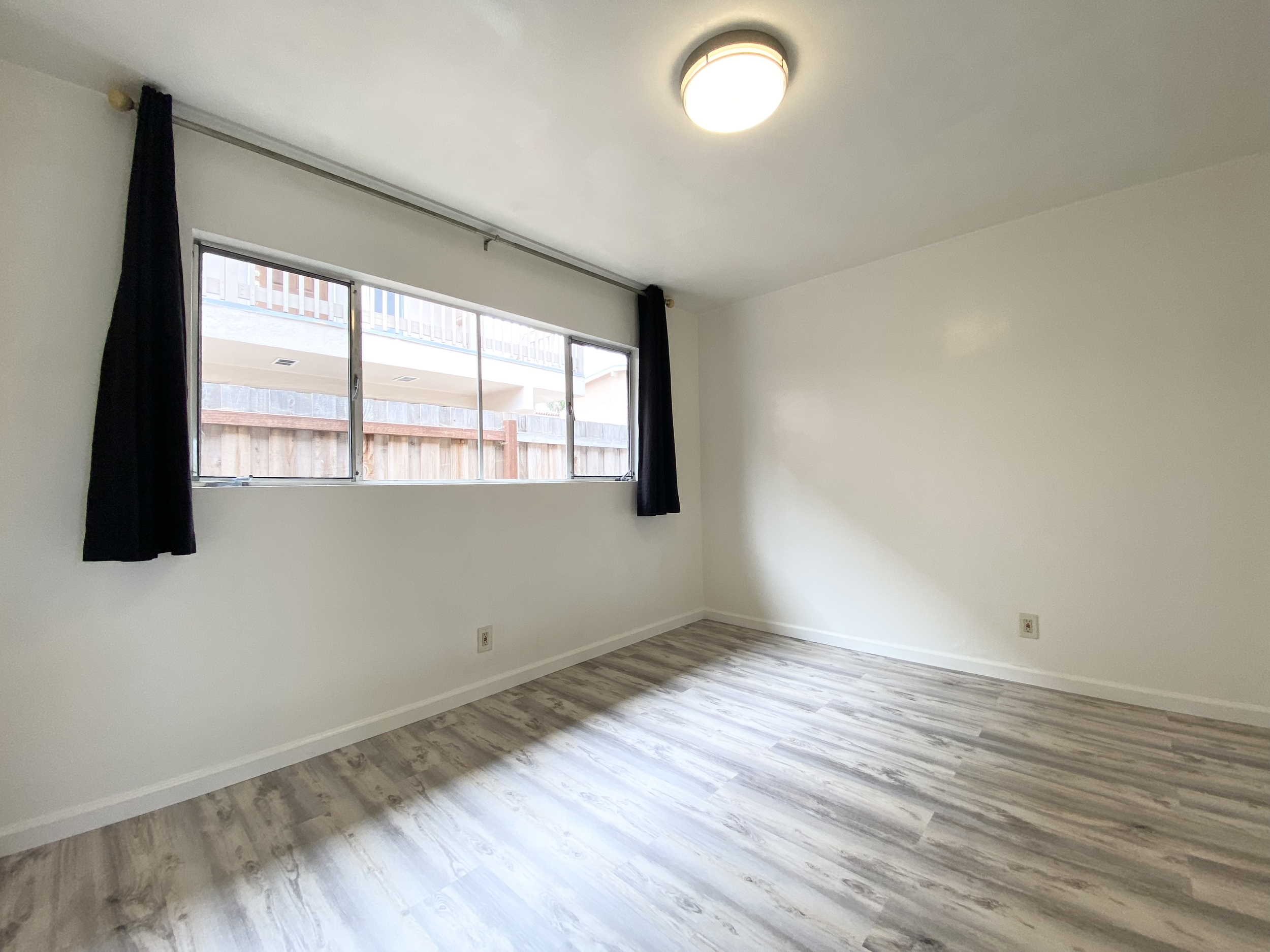Empty room with white walls, large window with black curtains, wood-like flooring, and ceiling light.