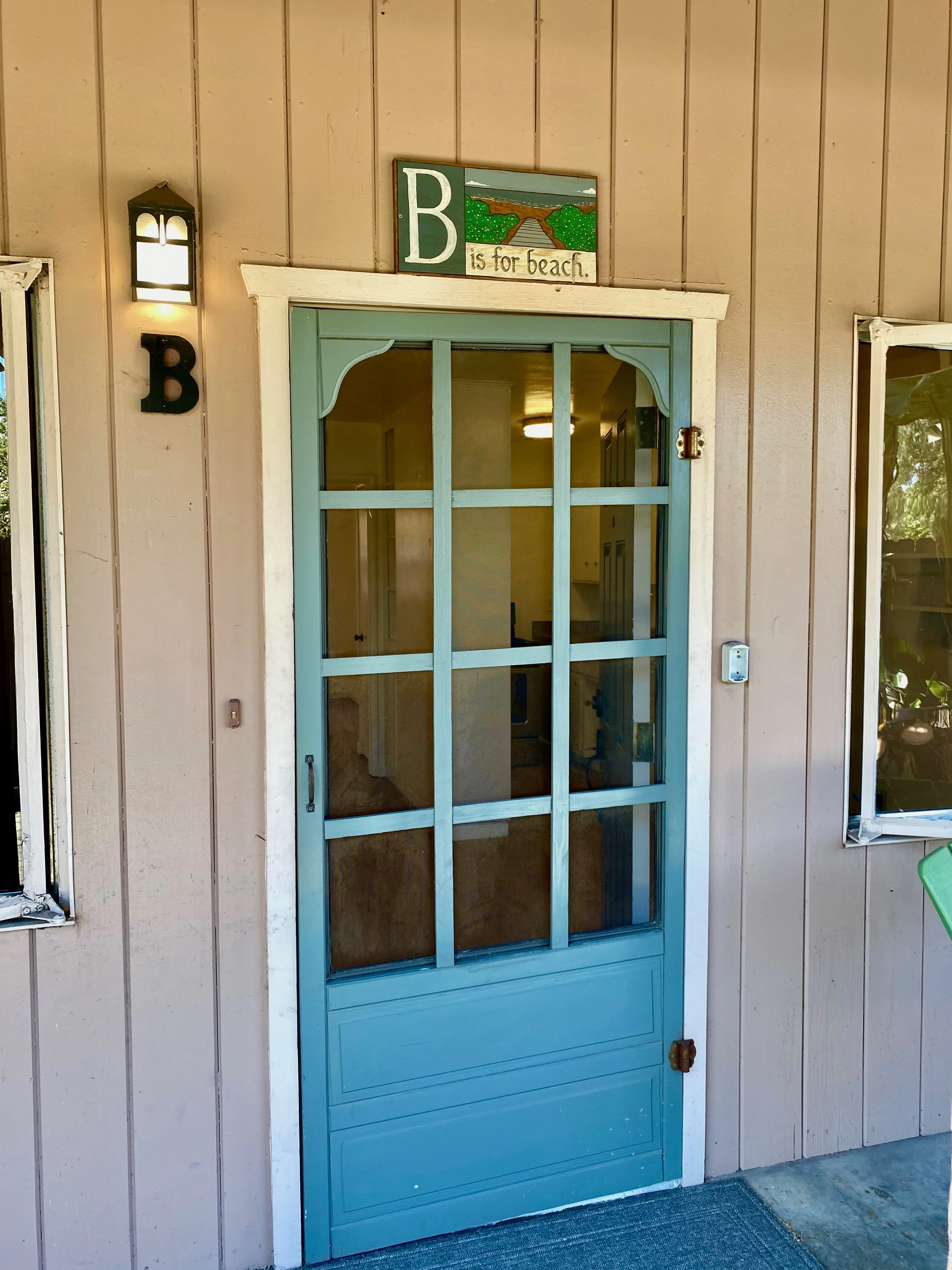 Light blue door with a screen door in front, mounted on a beige wooden siding building. A sign above the door reads 'B is for beach' with a painted scene of a beach and ocean.