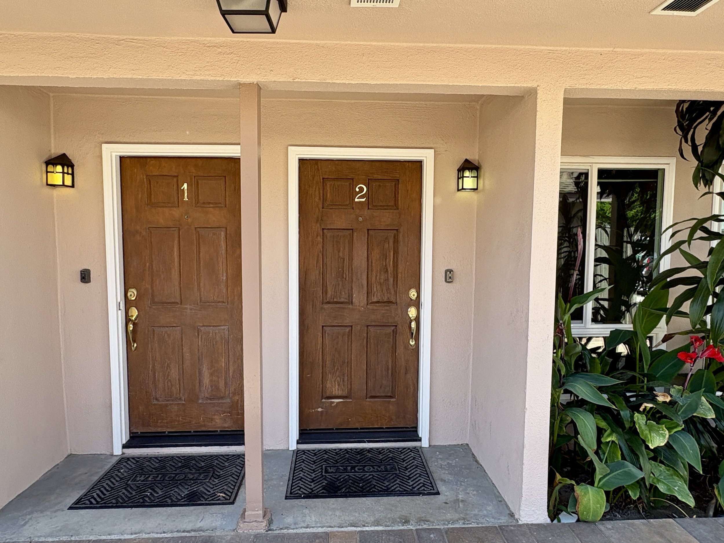 Double front doors made of wood with white trim, each door displaying a number 1 and 2, flanked by black lantern-style wall lights, with welcome mats in front and greenery to the right.