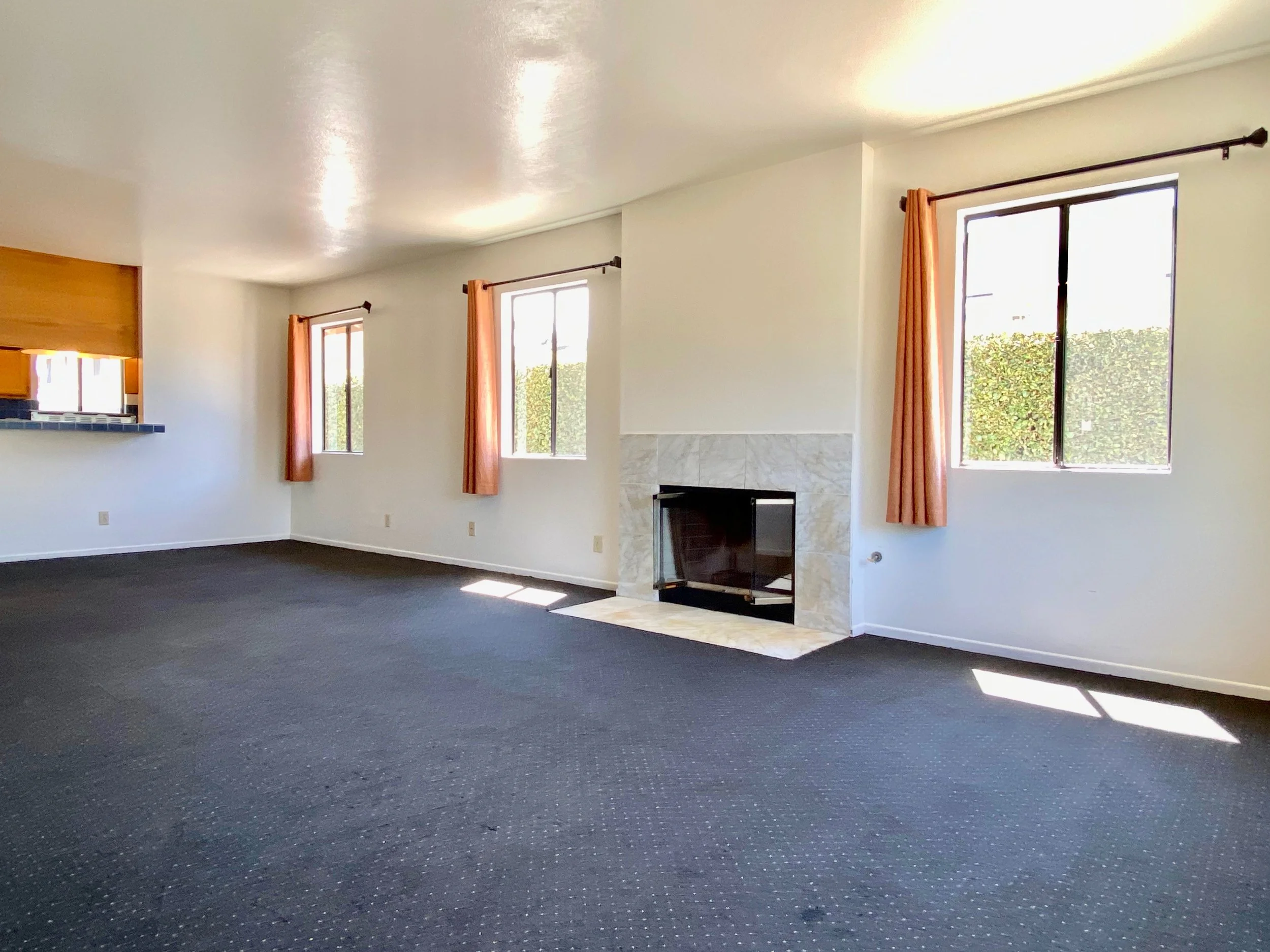 Empty living room with dark carpet, white walls, two windows with orange curtains, wood-trimmed doorway, and a fireplace with a marble surround.