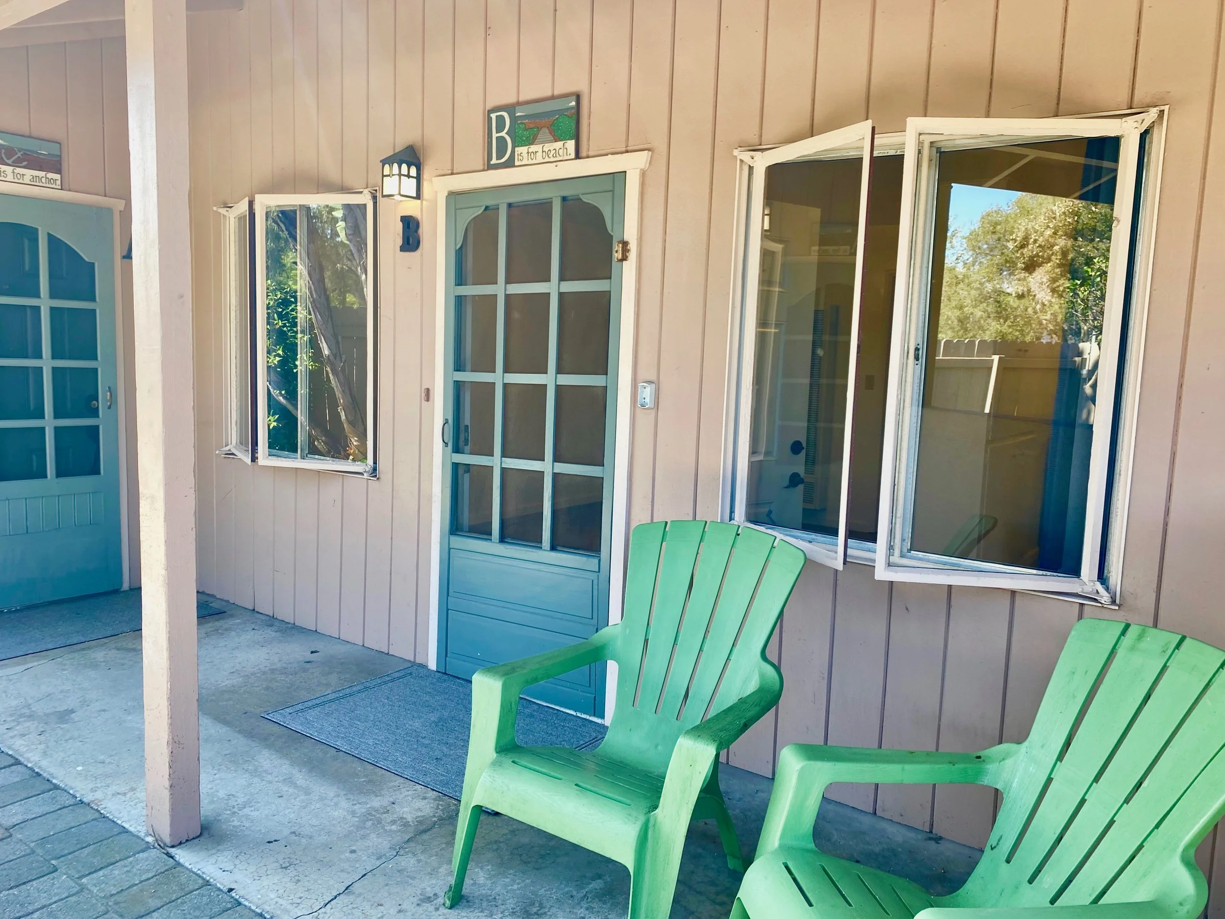 Green chairs outside a house with beige siding, blue door, and open window. A sign above the door says 'B is for beach' and there are decorative signs on the wall.