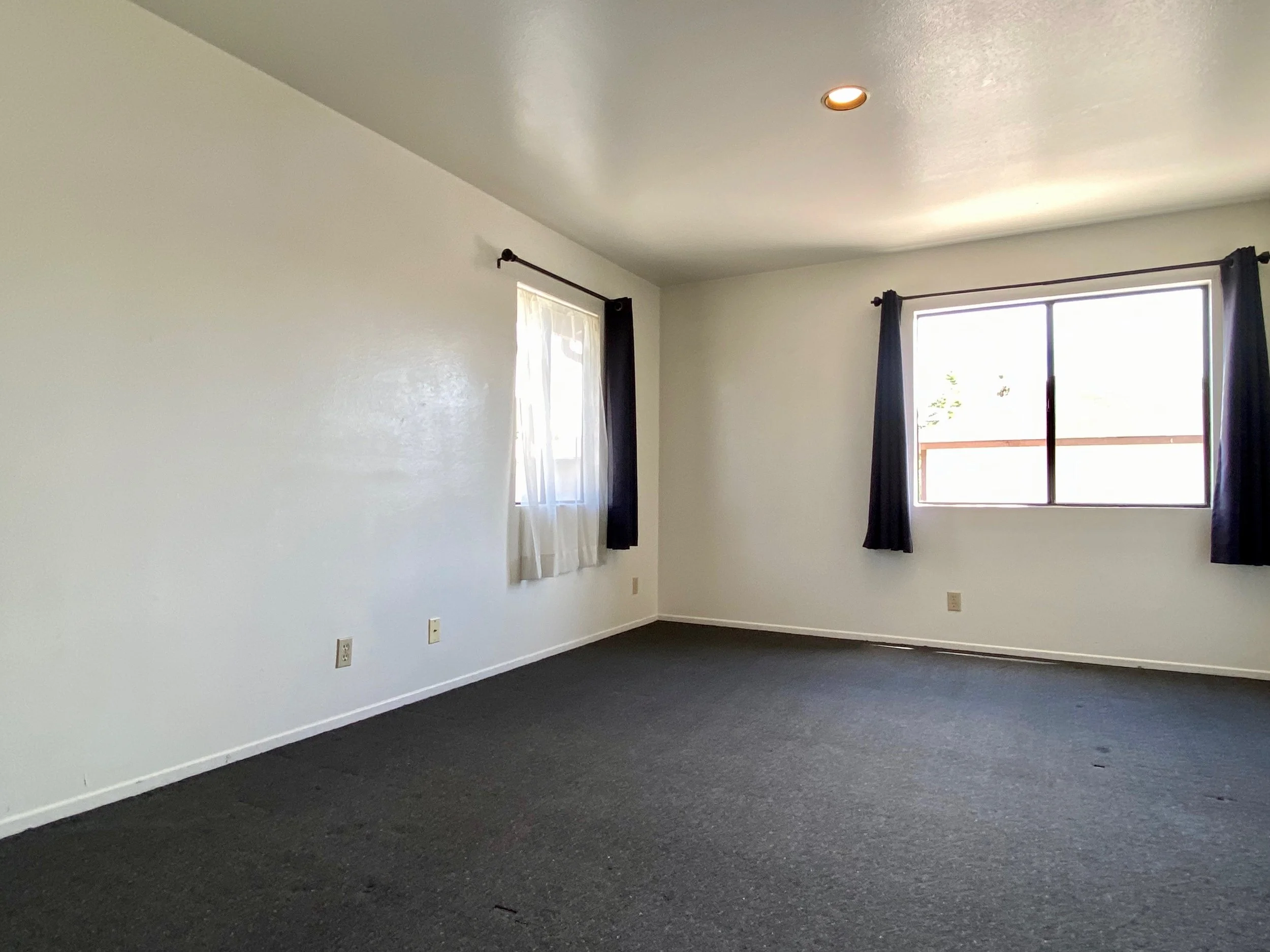 Empty room with white walls, two windows with dark curtains, and a dark carpeted floor.