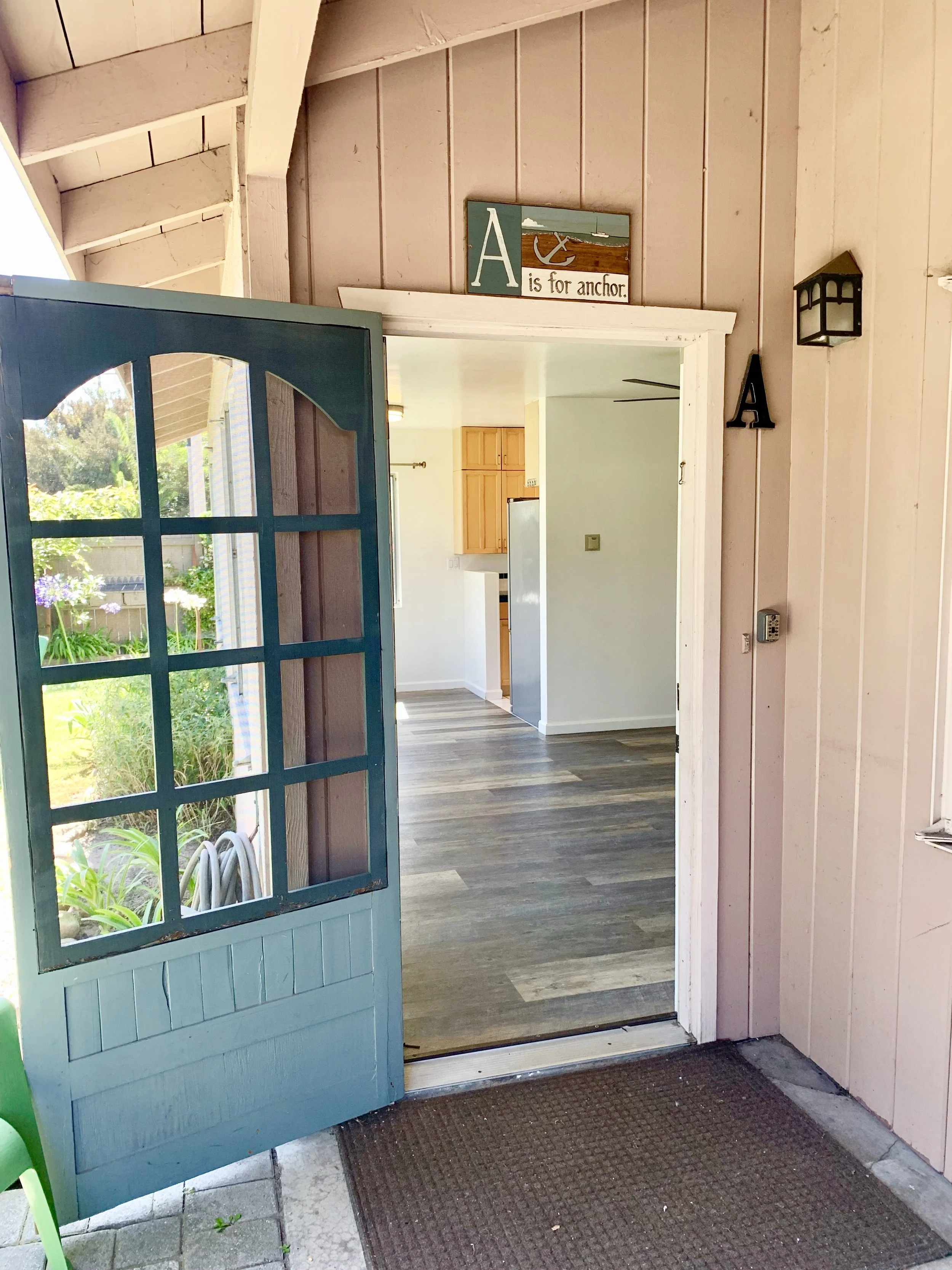 Open blue door leading from porch into a room with wood flooring and kitchen cabinets visible inside. A sign above the door reads 'A is for anchor,' with an illustration of an anchor.