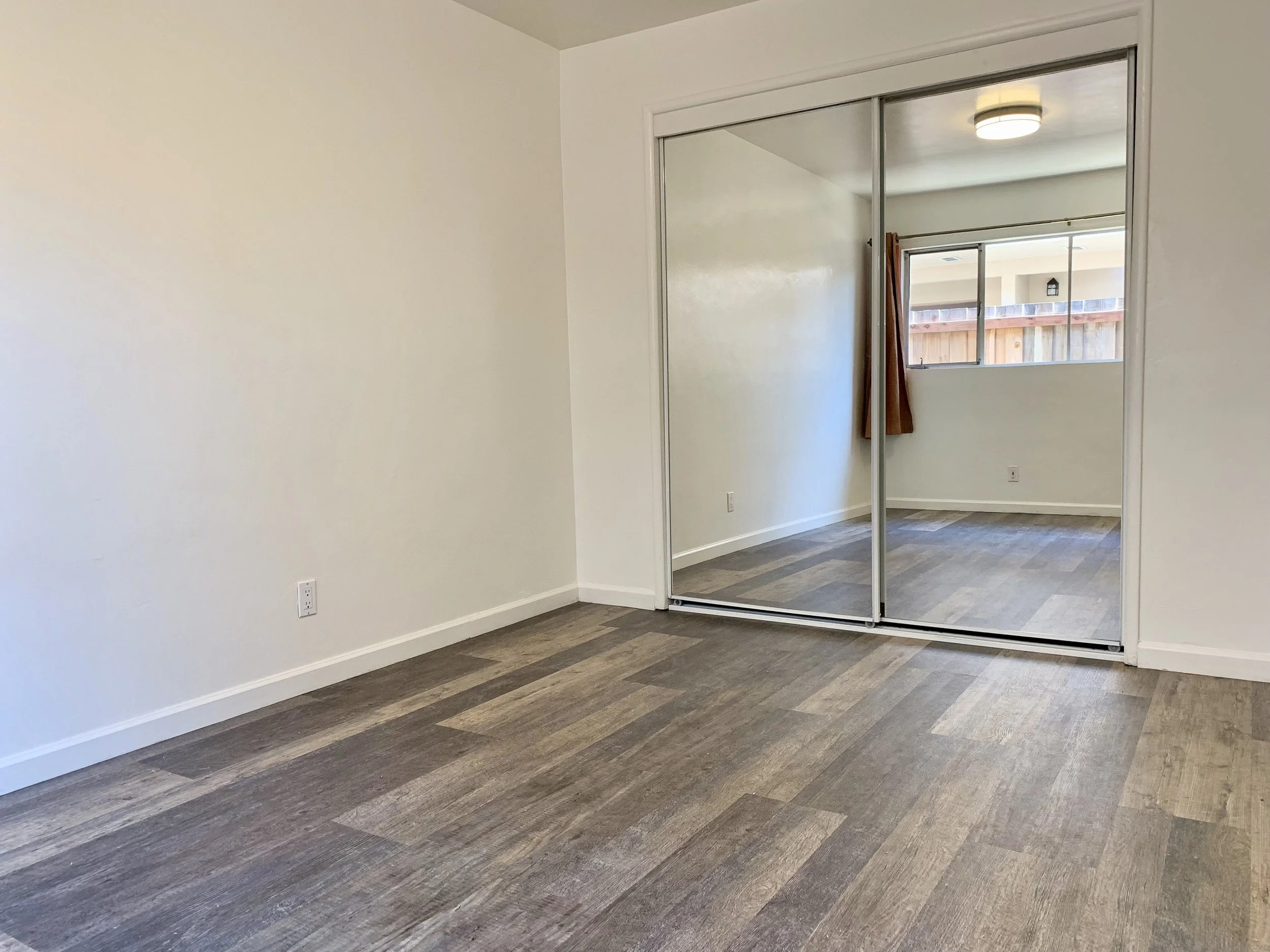 An empty room with white walls, hardwood flooring, a mirrored sliding closet door, and a window with a view of a wooden fence.