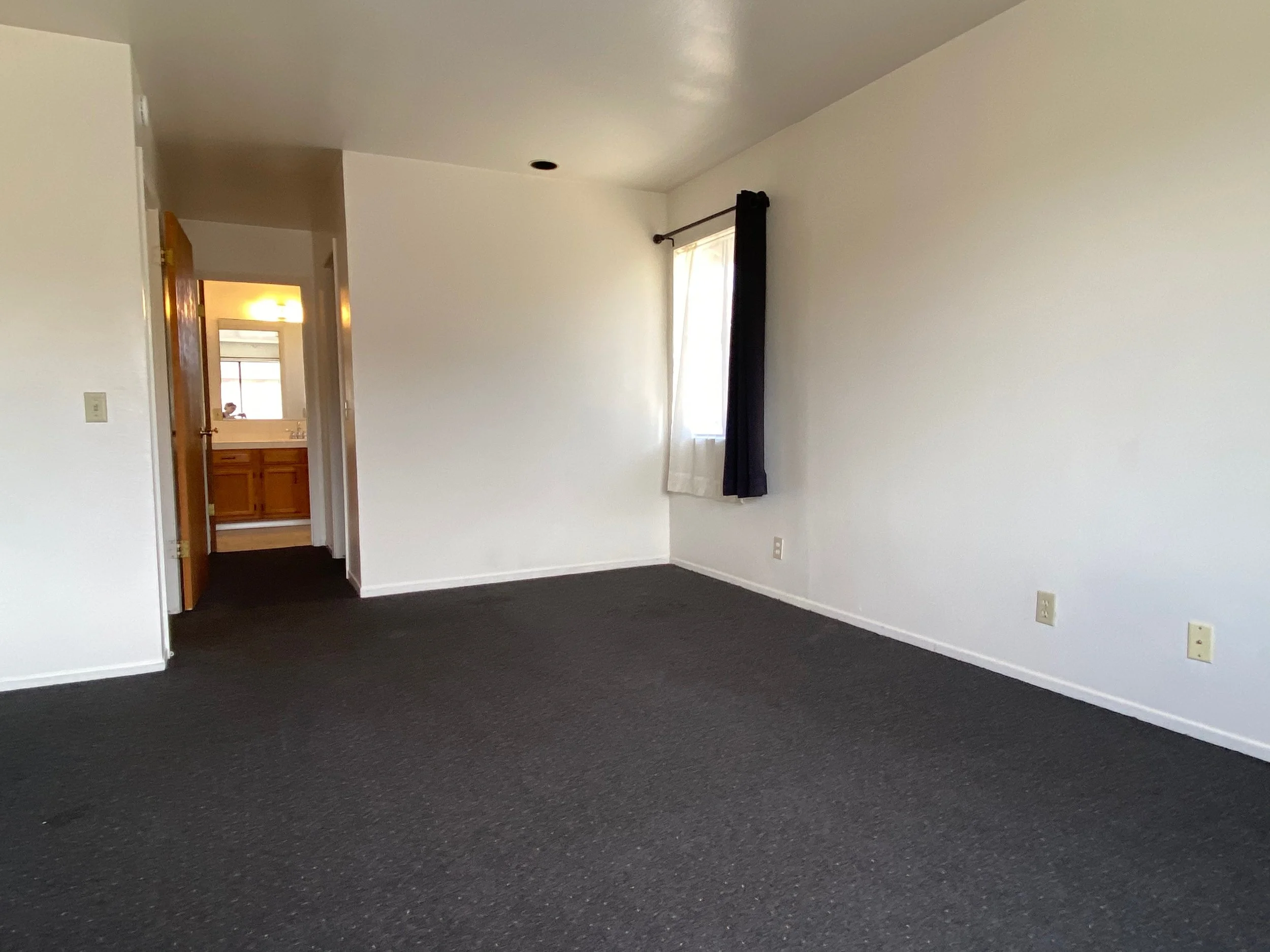 Empty room with dark carpet, white walls, a window with black curtains, and an open door leading to a bathroom with wooden cabinets and a mirror.