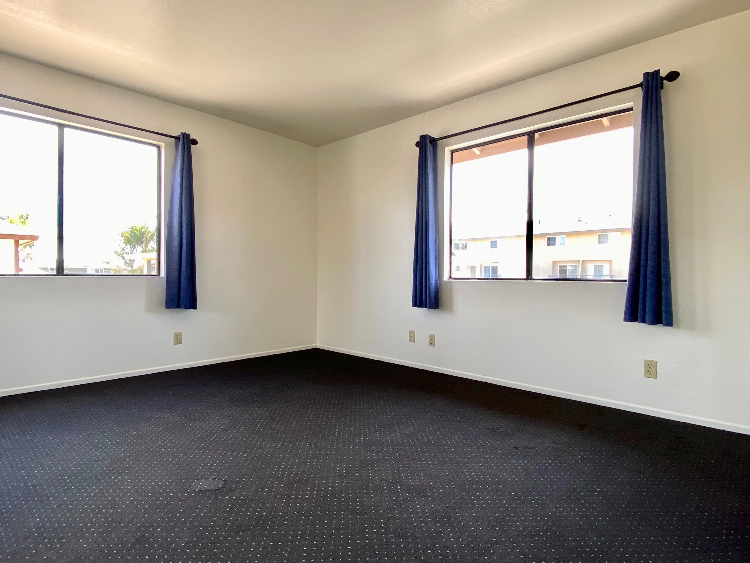 Empty room with three large windows, white-colored walls, blue curtains, dark patterned carpet, and some visible electrical outlets.