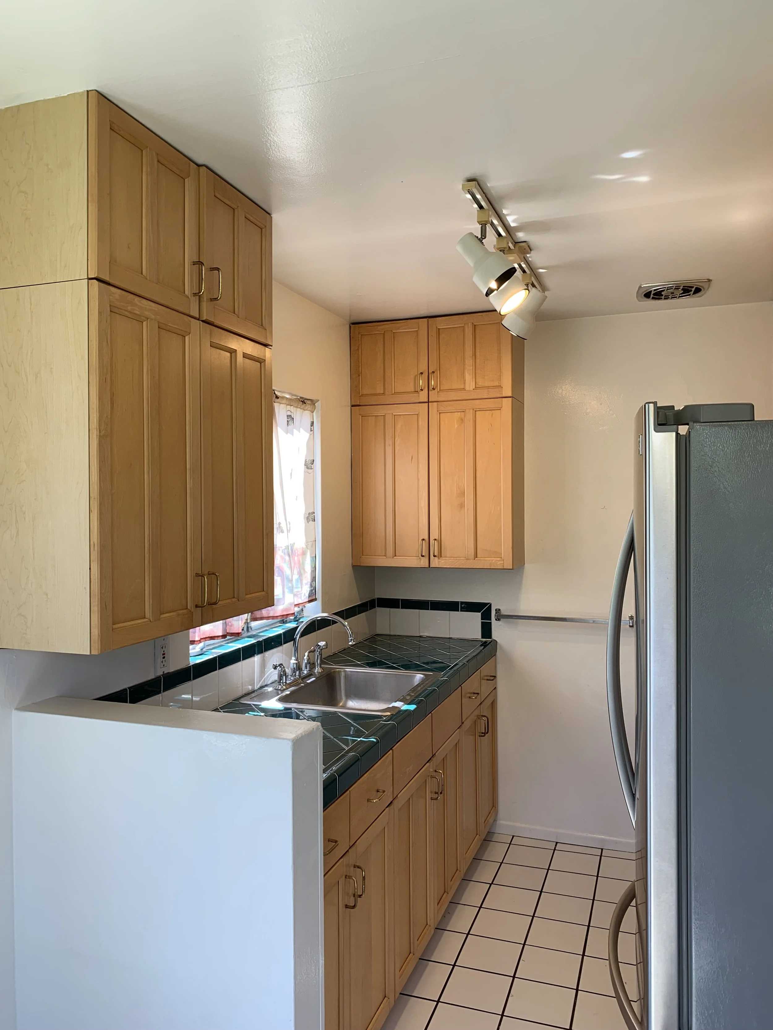A kitchen with wooden cabinets, a double sink, a tile countertop, a window with curtains, and a stainless steel refrigerator.