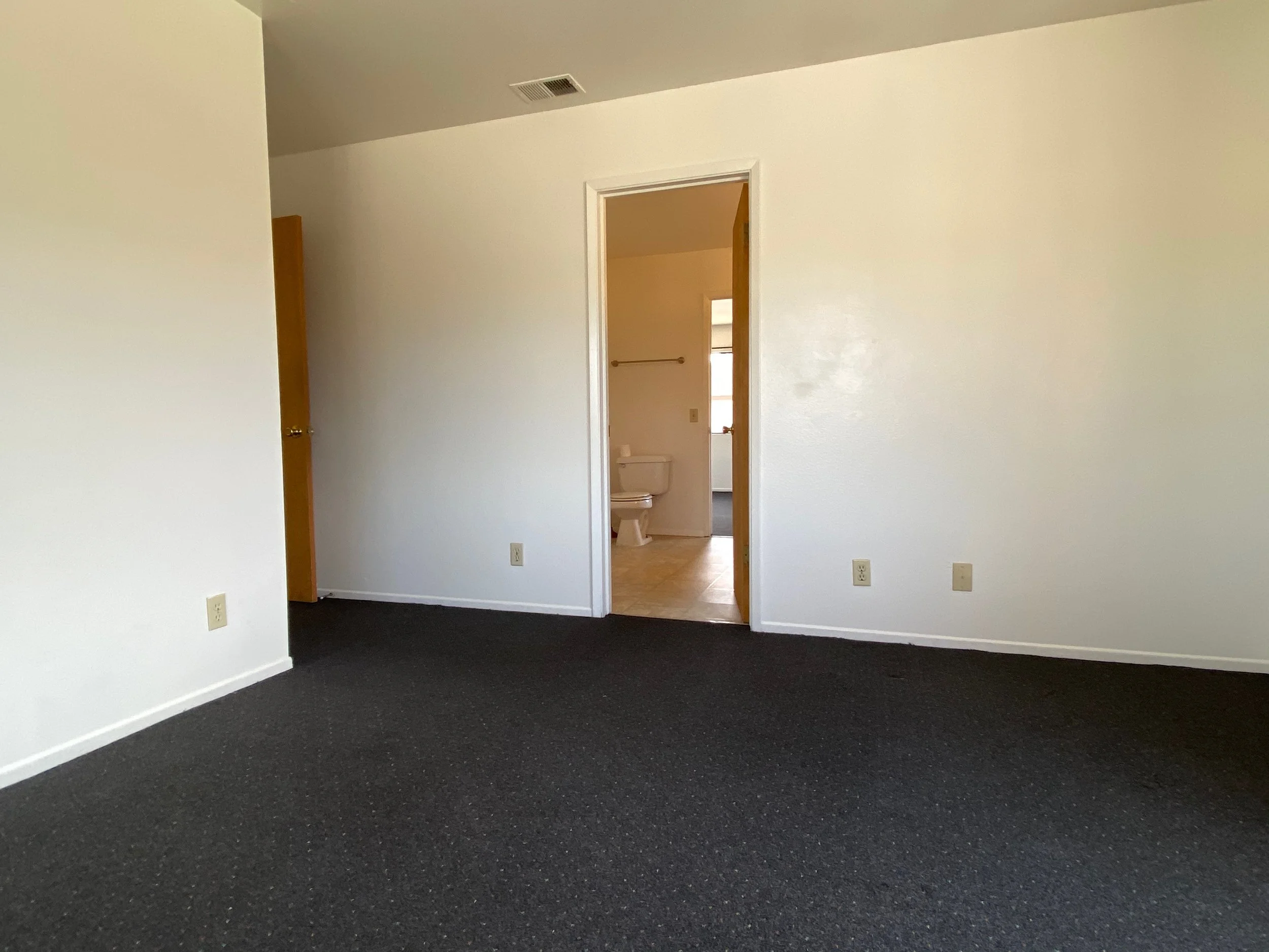 Empty room with black carpet, white walls, and a door leading to a bathroom with a toilet. There are electrical outlets on the walls and a window visible in the bathroom.