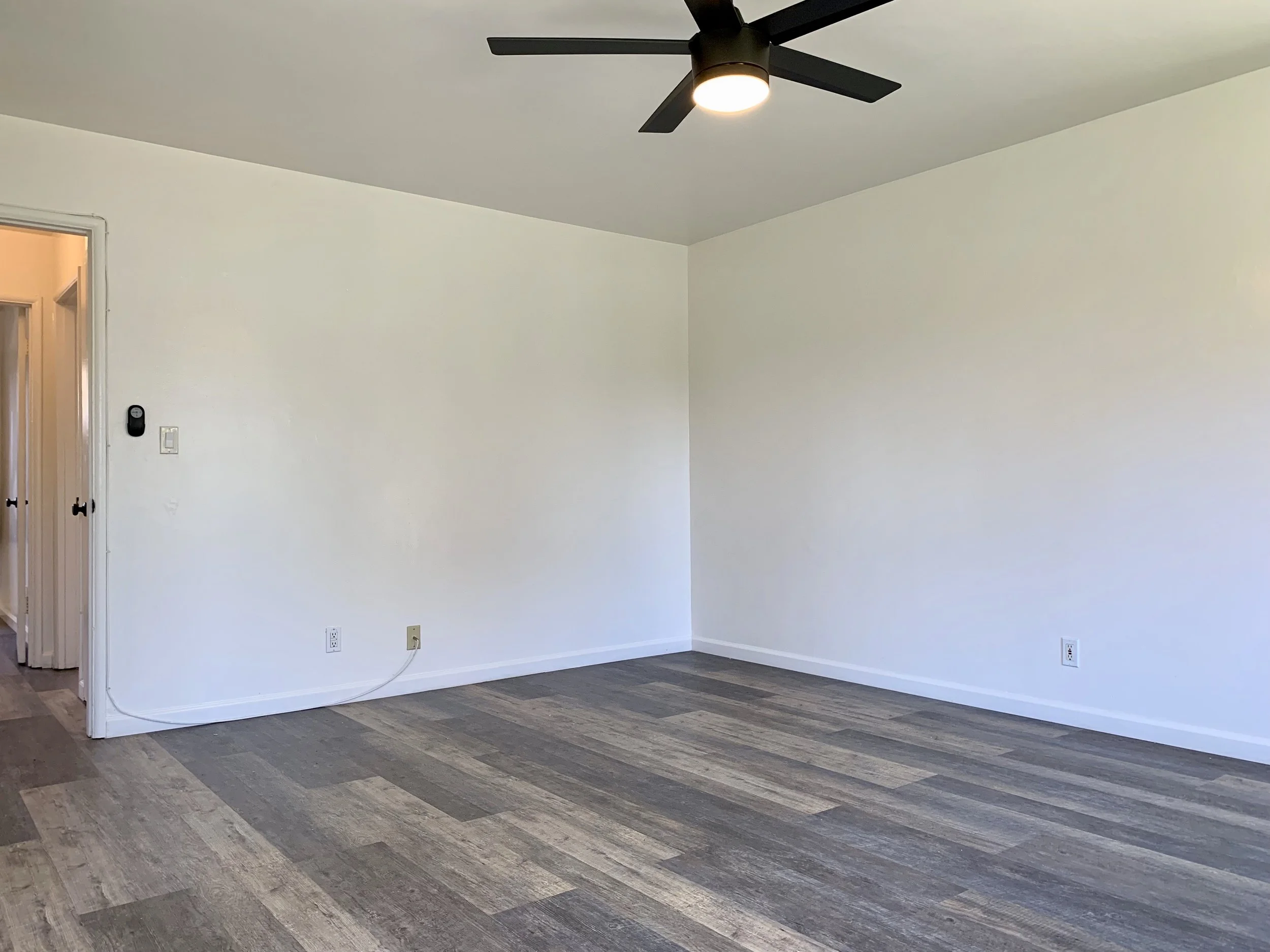 Empty room with white walls, ceiling fan with light, wood-like flooring, and a doorway on the left showing an adjacent room with a light-colored wall.