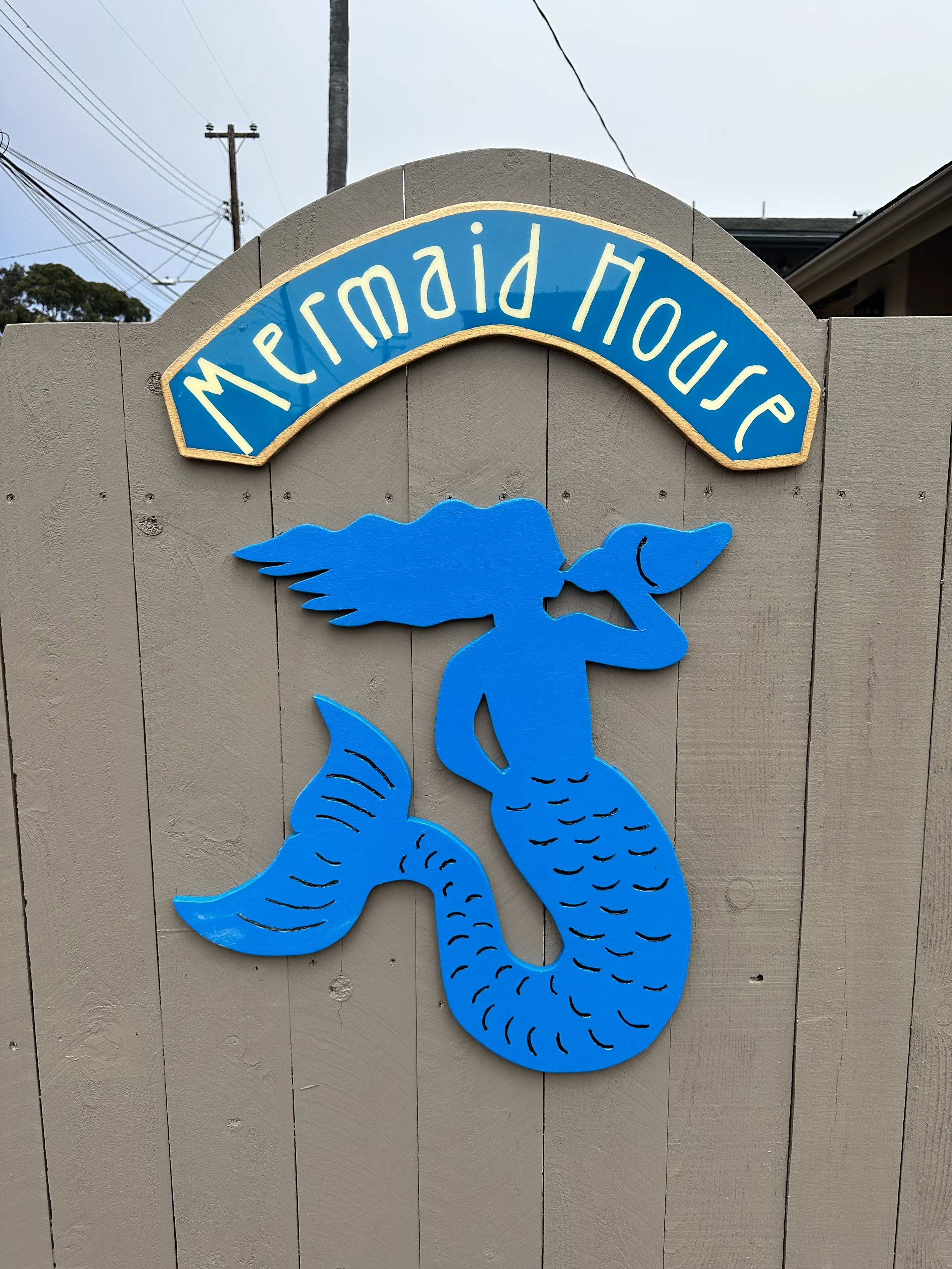 Decorative sign with a blue mermaid holding a conch shell and a sign that reads 'Mermaid House' in yellow letters.