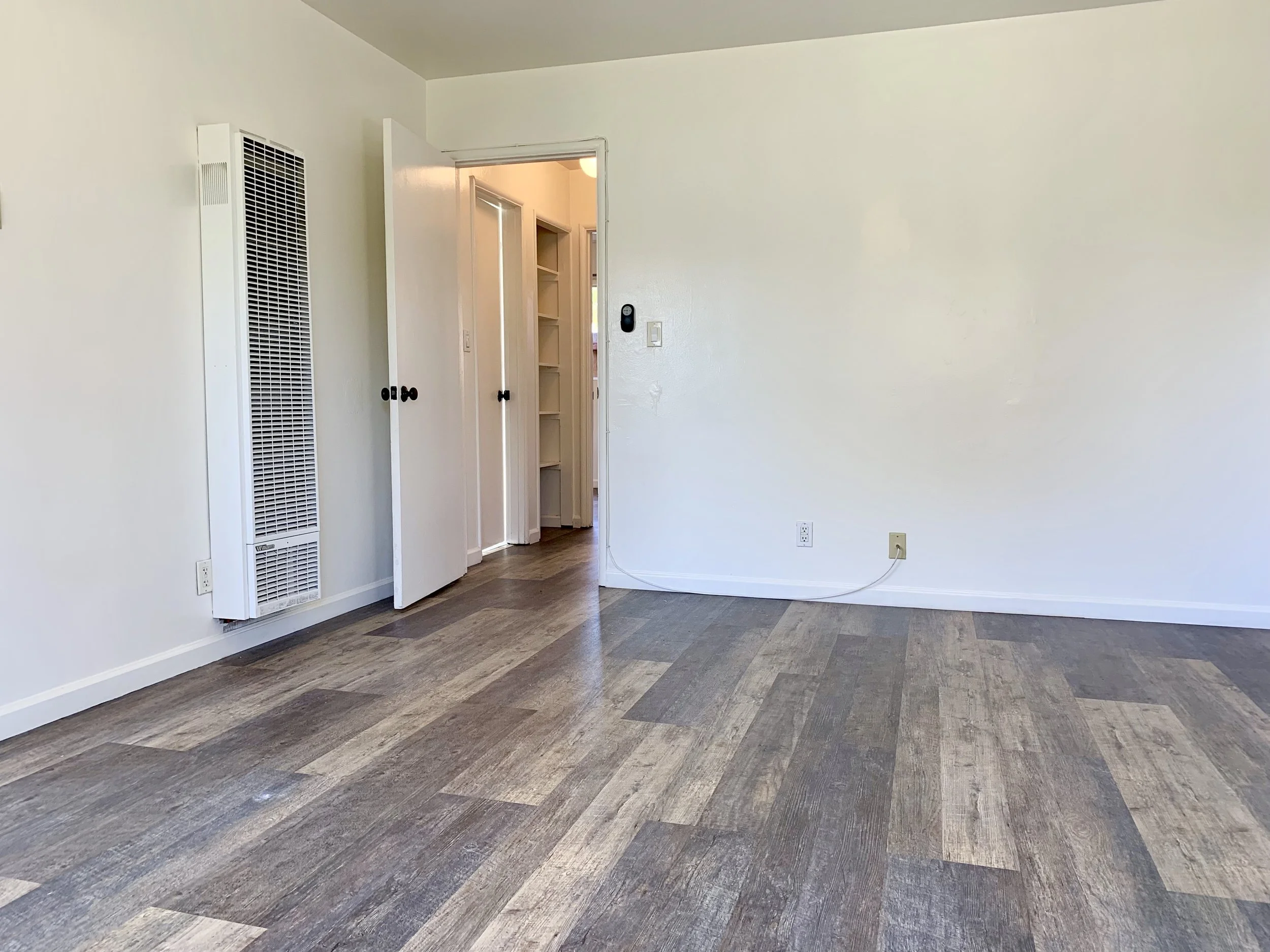 Empty room with white walls, a door open to hallway, wooden floor, and wall heater.