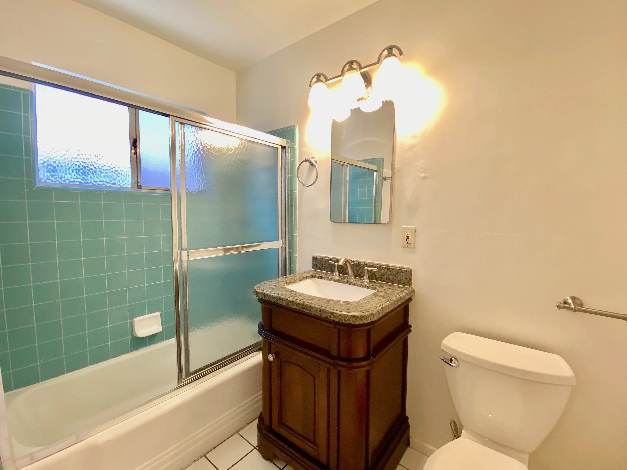 Bathroom with green tiled shower walls, a shower with frosted glass doors, a mirror above a granite countertop, and a toilet.
