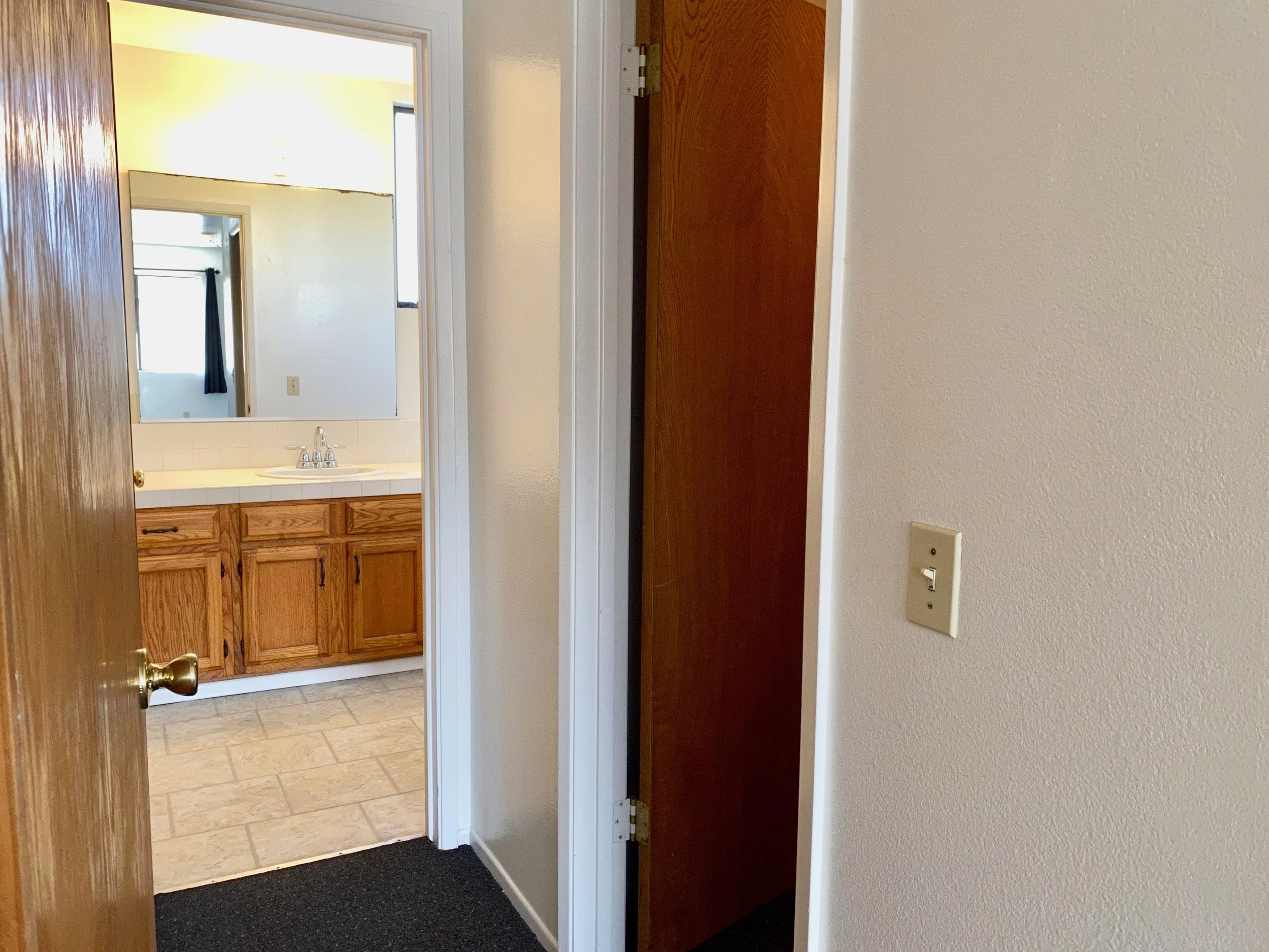 View of a bathroom through an open door from a hallway. The bathroom has a wooden cabinet, a sink, tiled flooring, and a window with a curtain.