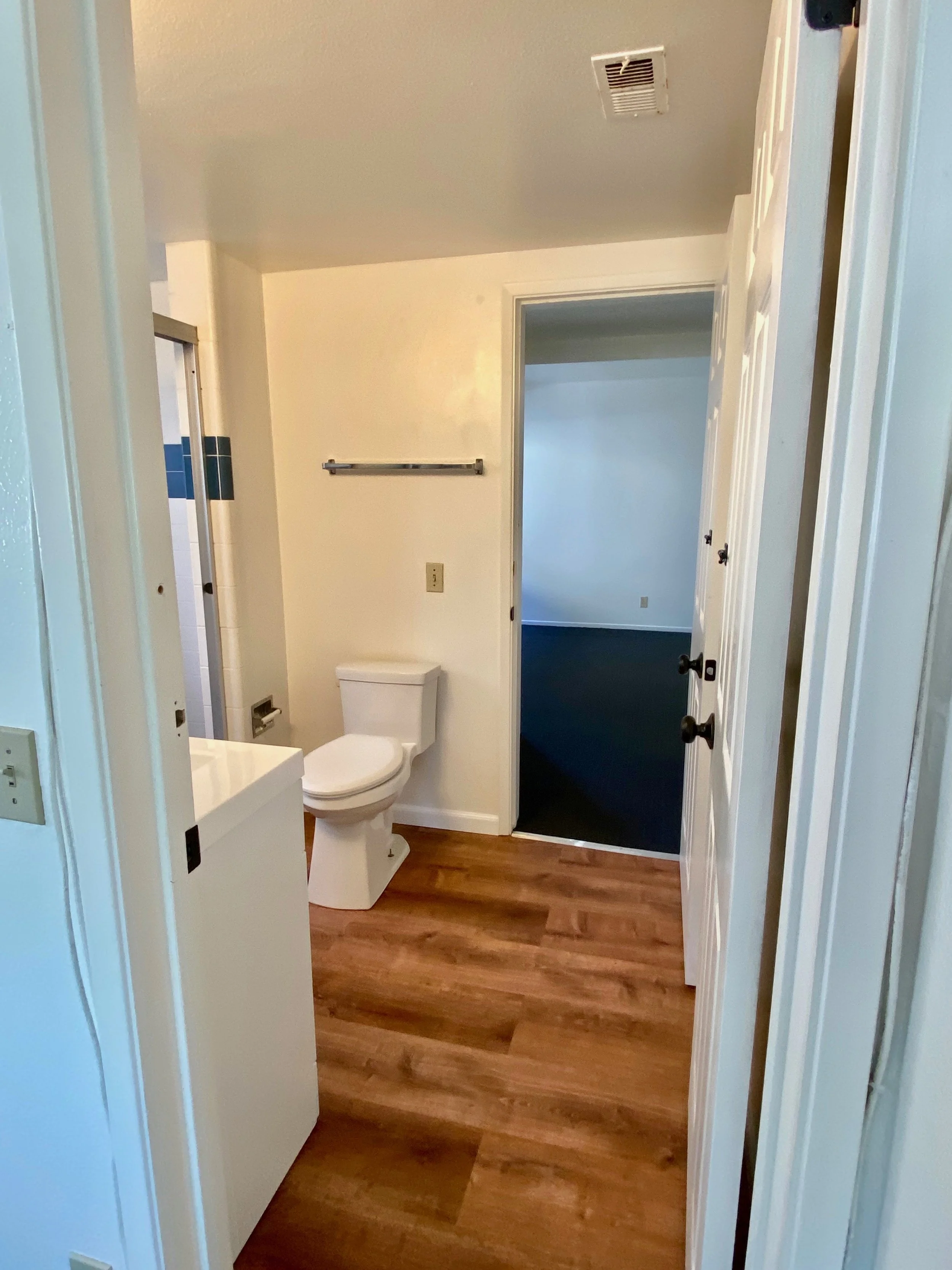 A small bathroom with a toilet, hardwood flooring, and a doorway leading to another room painted in dark blue. The bathroom has off-white walls, a vent on the ceiling, and some cabinetry or fixtures partially visible on the right side.