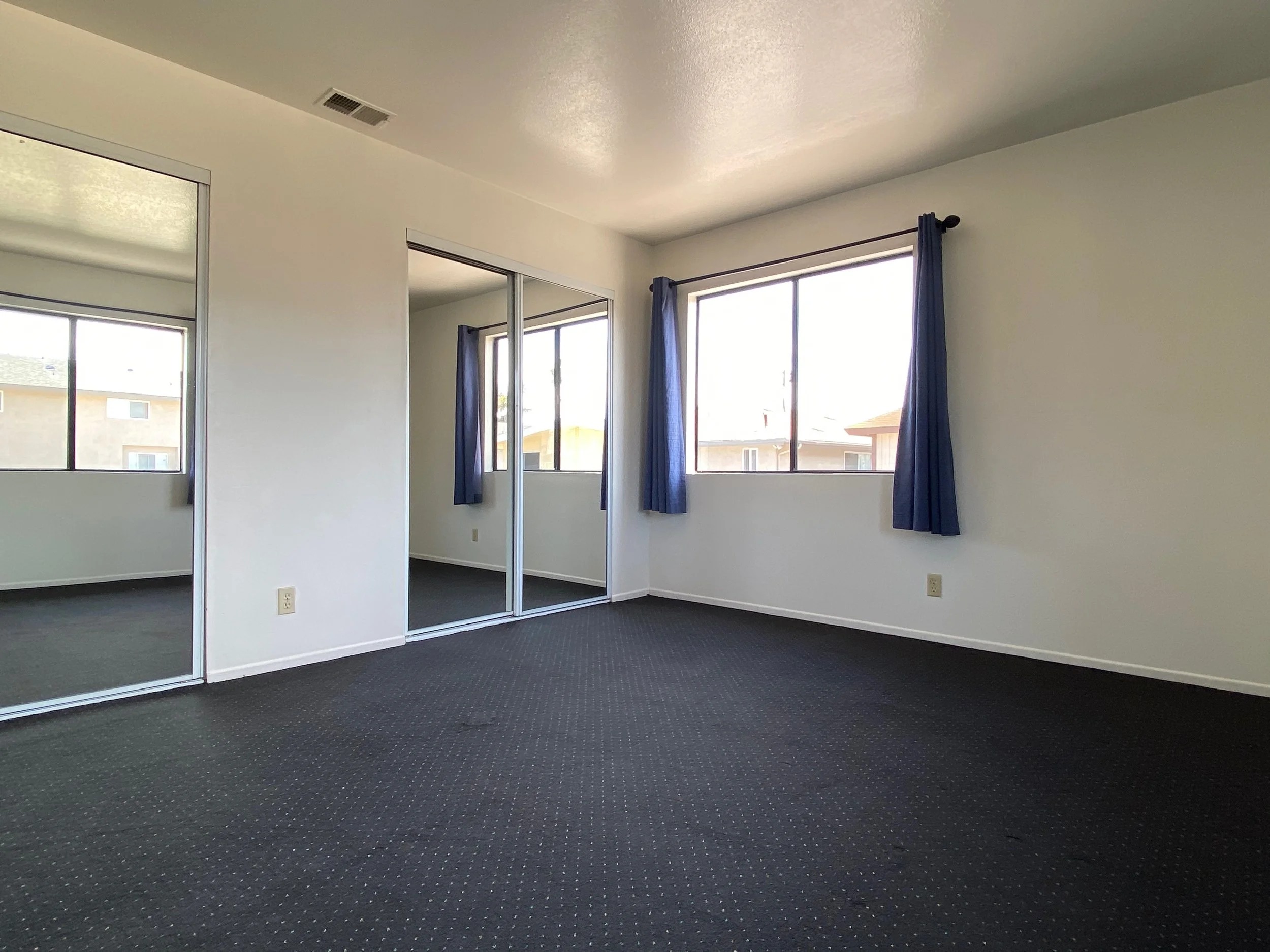 Empty room with large windows, dark curtains, mirrored closet doors, and dark carpet flooring.