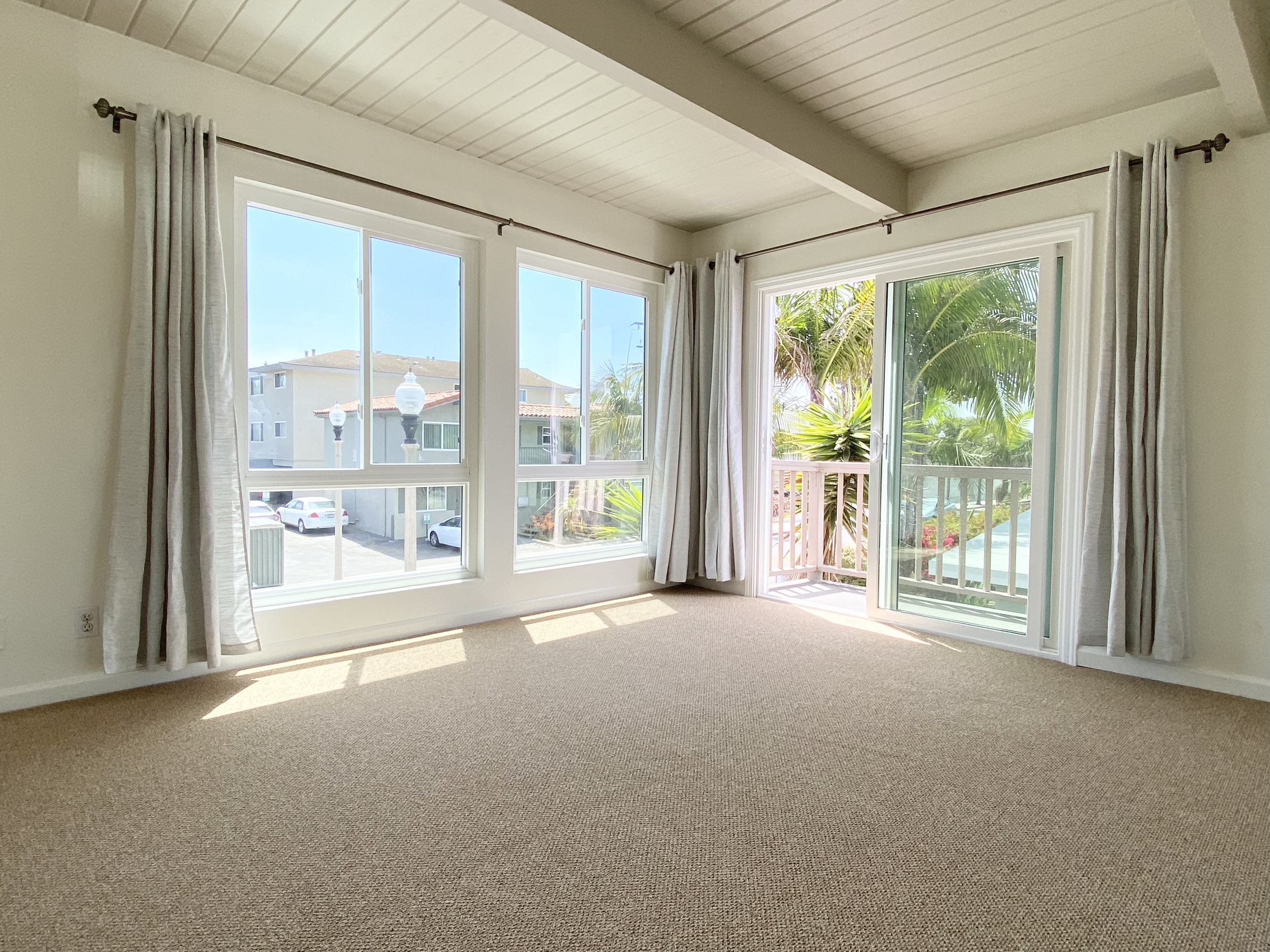 A living room with beige carpet, large windows with white curtains, and a sliding glass door leading to a balcony with green palm trees outside. The ceiling is paneled and there are no furniture or decorations in the room.