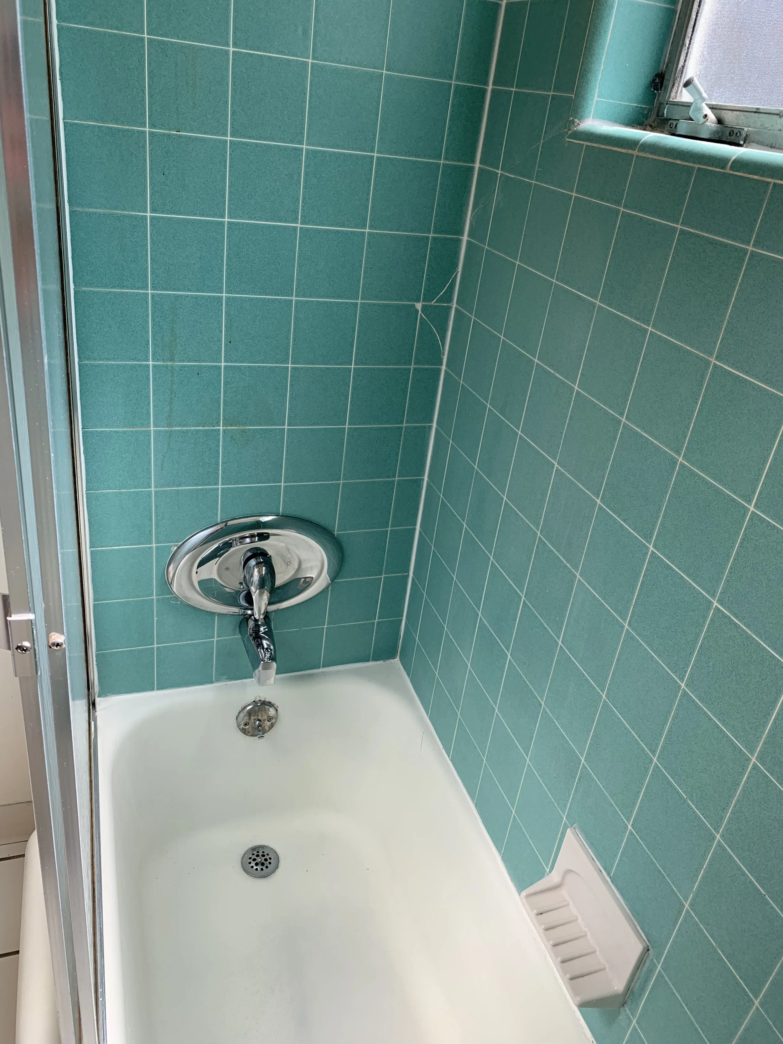 A shower bathroom with green tile walls, a white shower base, a chrome shower drain, and a chrome shower faucet. There is a small window with a screen and a metal latch in the upper right corner.