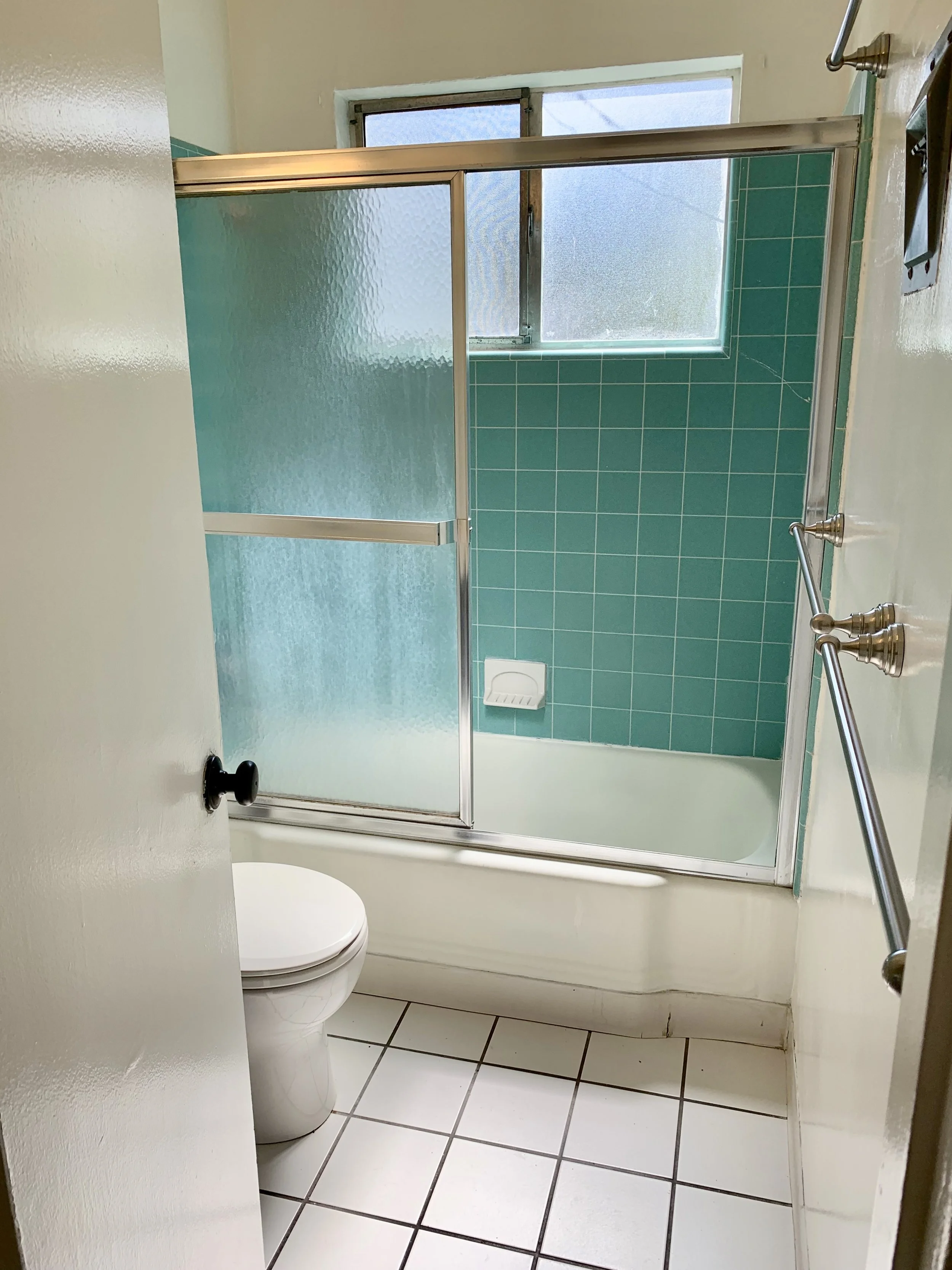 A small bathroom with a white toilet, a walk-in shower with frosted glass door, teal wall tiles, and a window above the shower.
