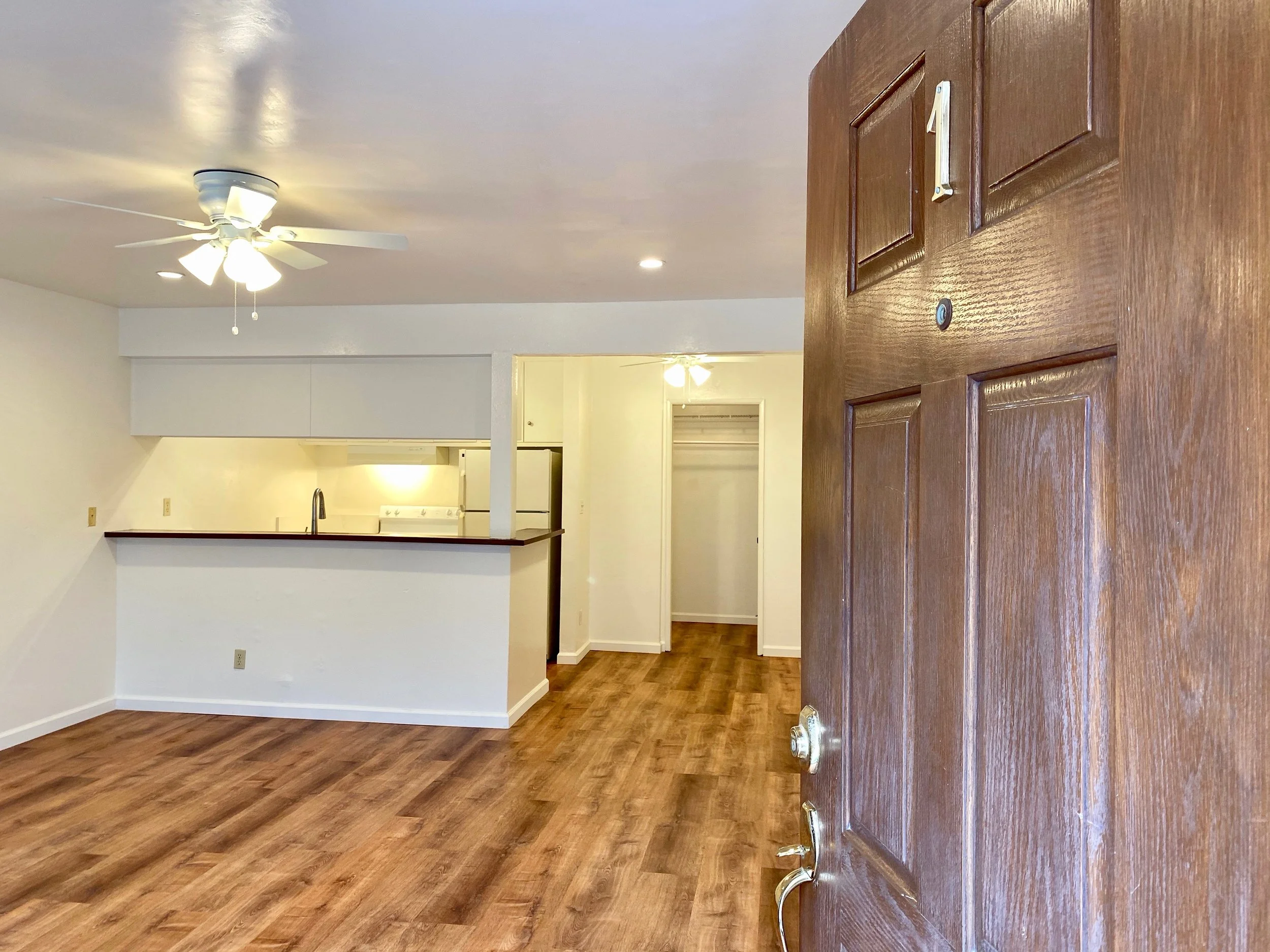View of a room with wood flooring, a white ceiling fan, and an open kitchen area in the background. 