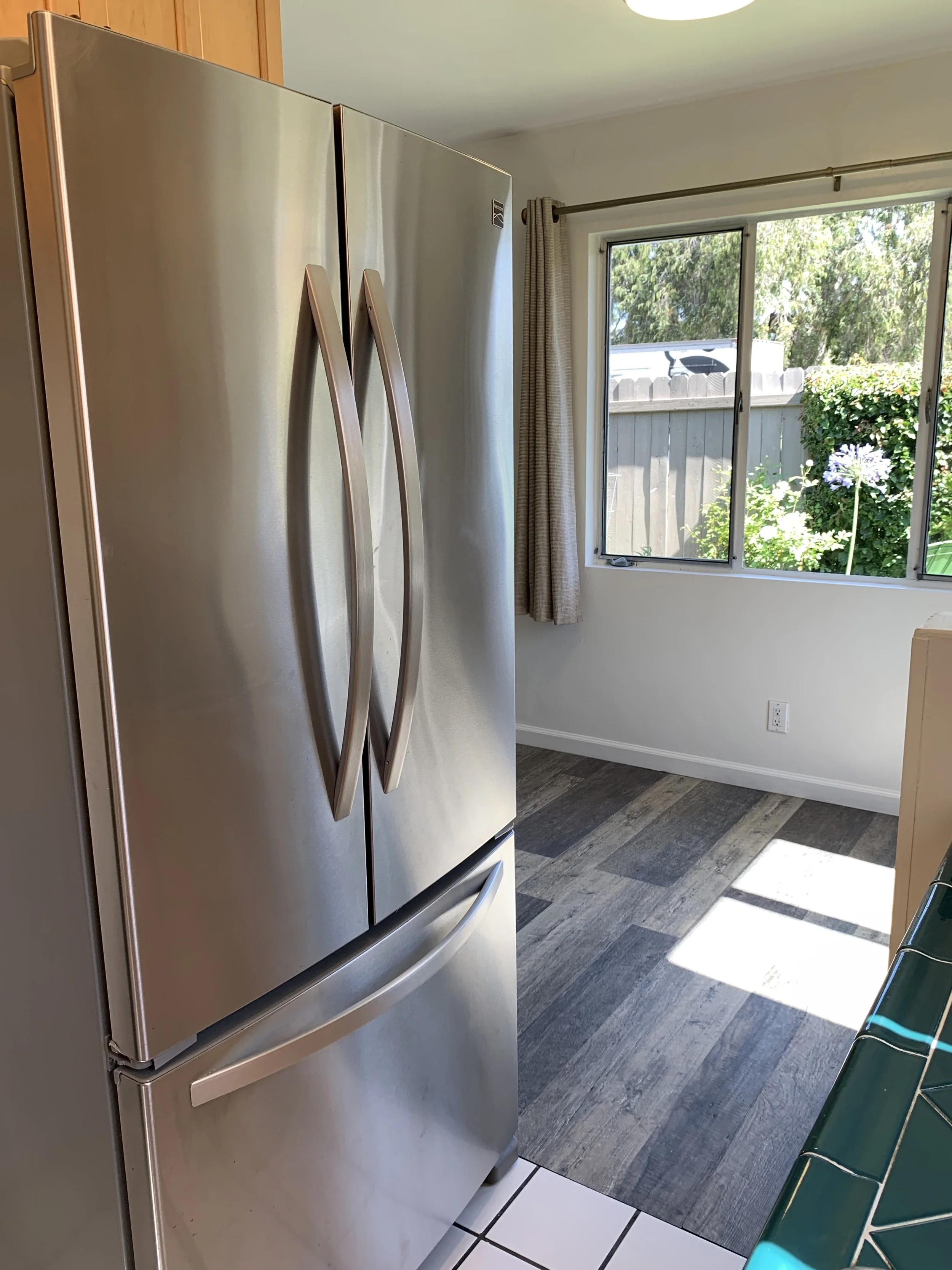 Stainless steel refrigerator with double doors in a kitchen next to a window with beige curtains, a garden with greenery and flowers outside.