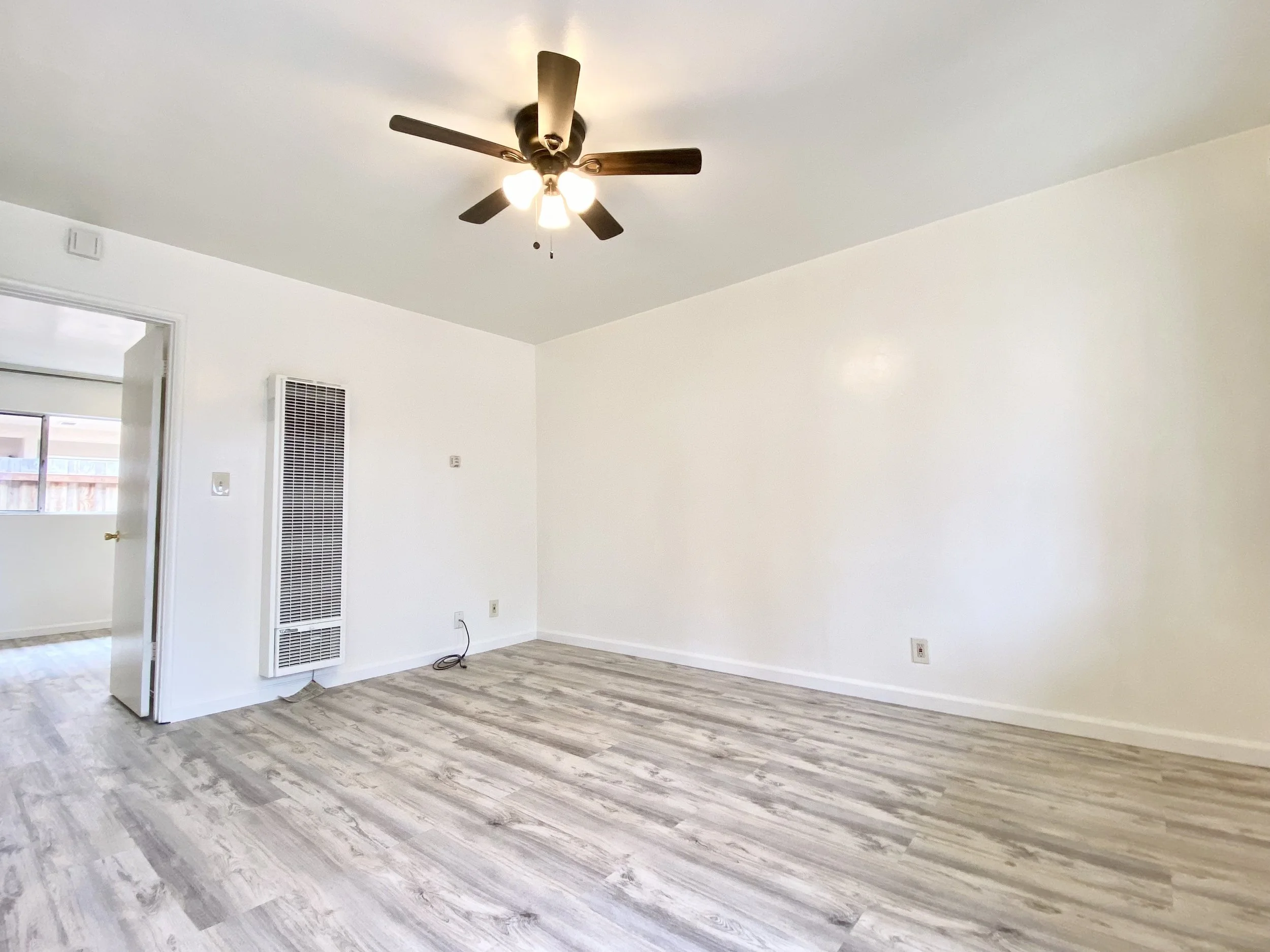 An empty living room with white walls, a ceiling fan with lights, wooden flooring, and a doorway leading to another room with a window.