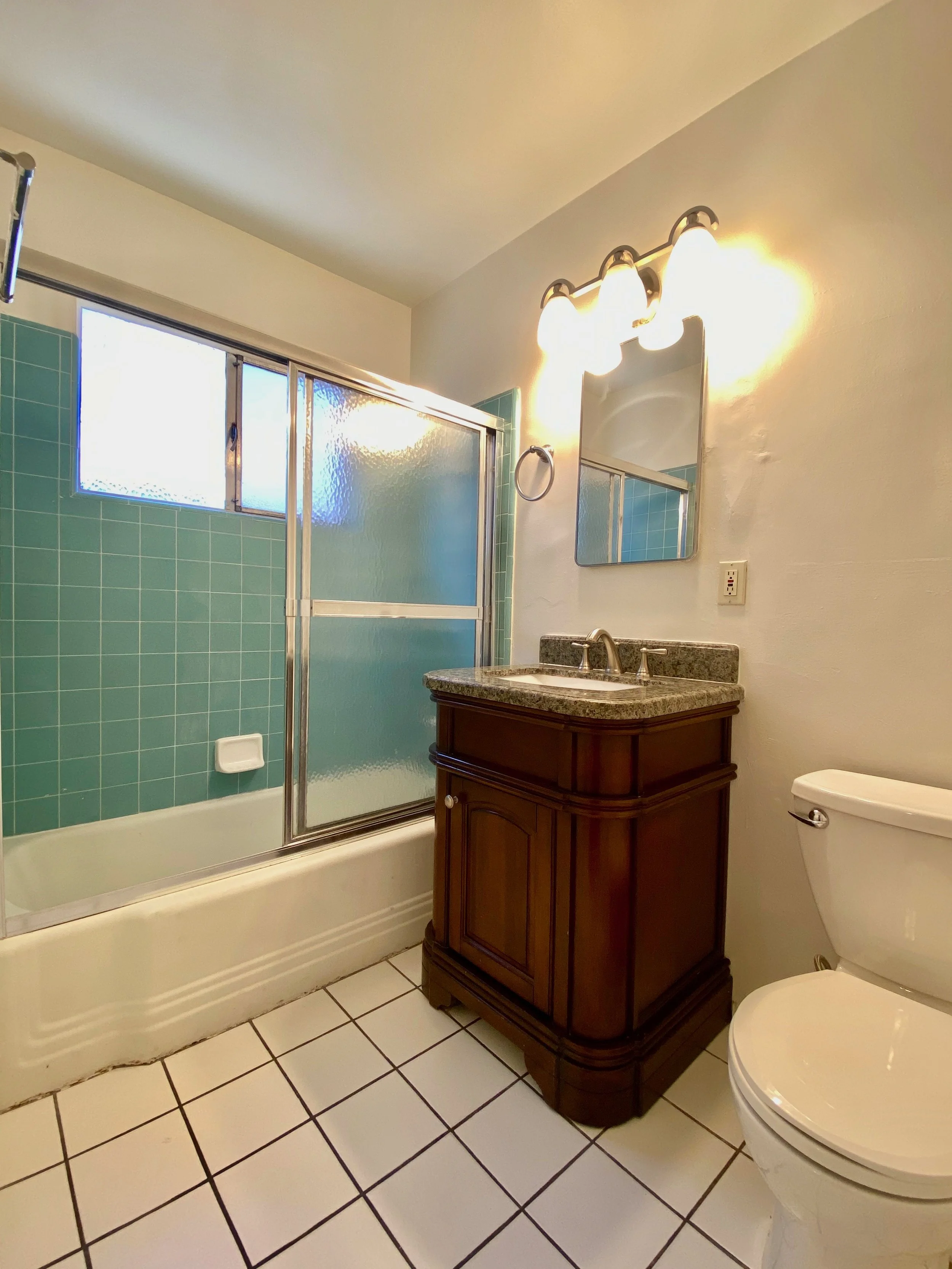 A bathroom featuring a shower with a frosted glass sliding door and teal tiles, a wooden vanity with a granite countertop, a mirror, a light fixture, and a toilet on white tiled floor.