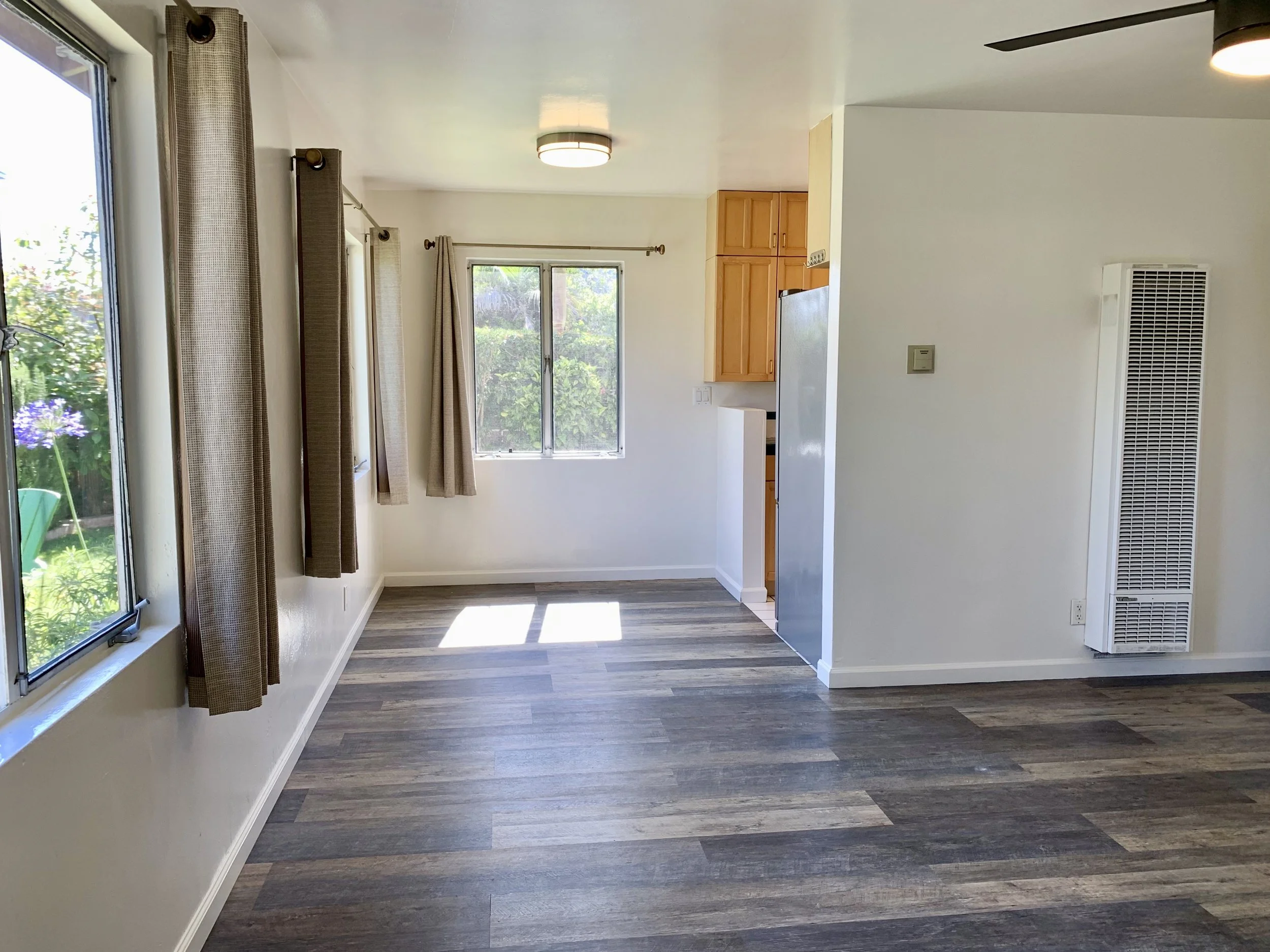Bright and spacious living room with large windows, curtains, wood flooring, and a ceiling light. Part of the kitchen with wooden cabinets and a refrigerator is visible.