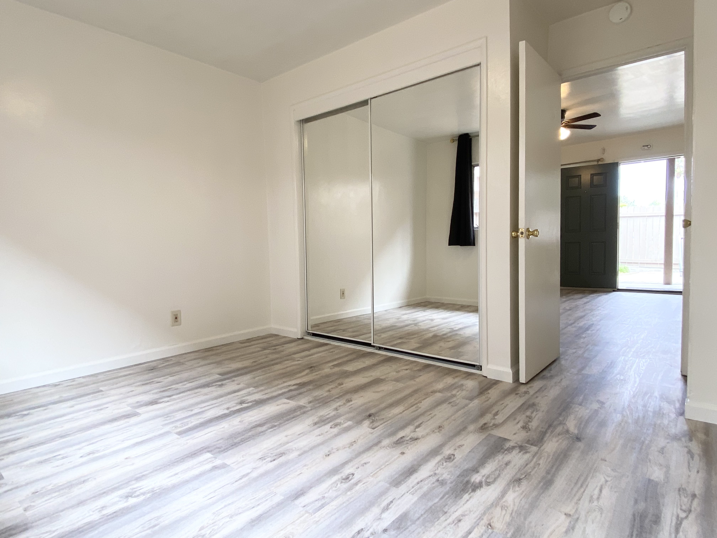 Empty room with wood-patterned flooring, a built-in wardrobe with mirrored sliding doors, and an open door leading to another room with a dark green door and a ceiling fan