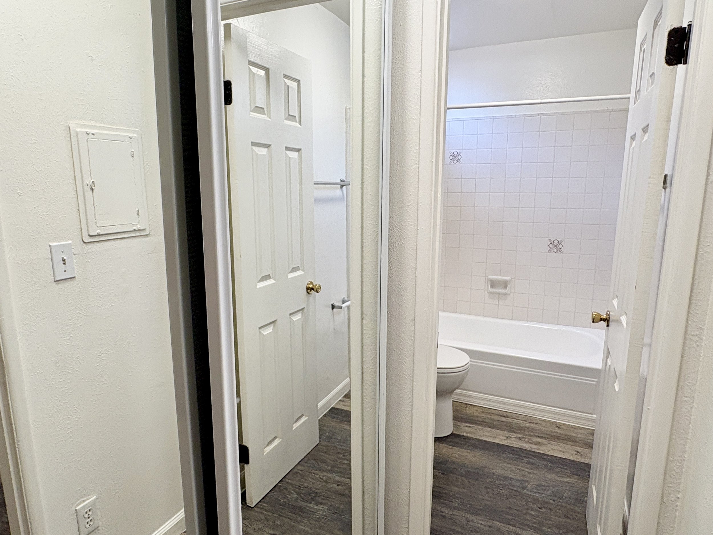 View of a small bathroom with a bathtub and toilet, seen through a doorway. The bathroom has white tiled walls and dark wood floor.
