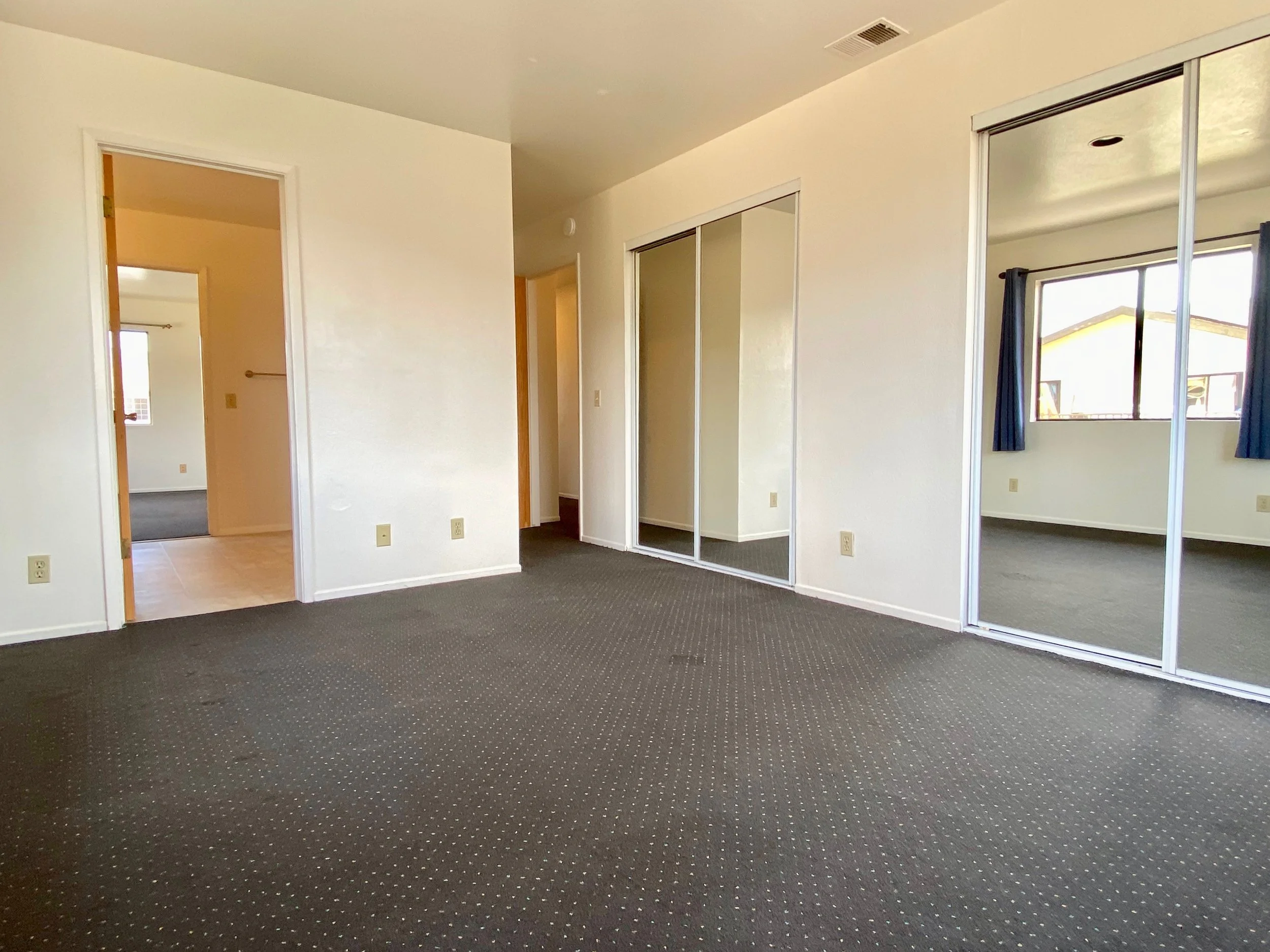 Room with dark carpeted flooring, white walls, large windows with curtains, closet with mirrored sliding doors, and doorways leading to a bathroom and hallway.