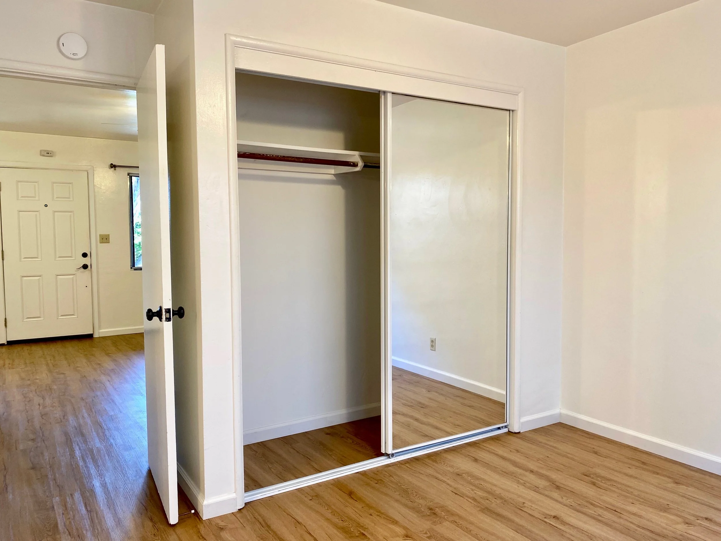 A bedroom with a closet that has a sliding mirror door, hardwood floors, a white door, and a window.