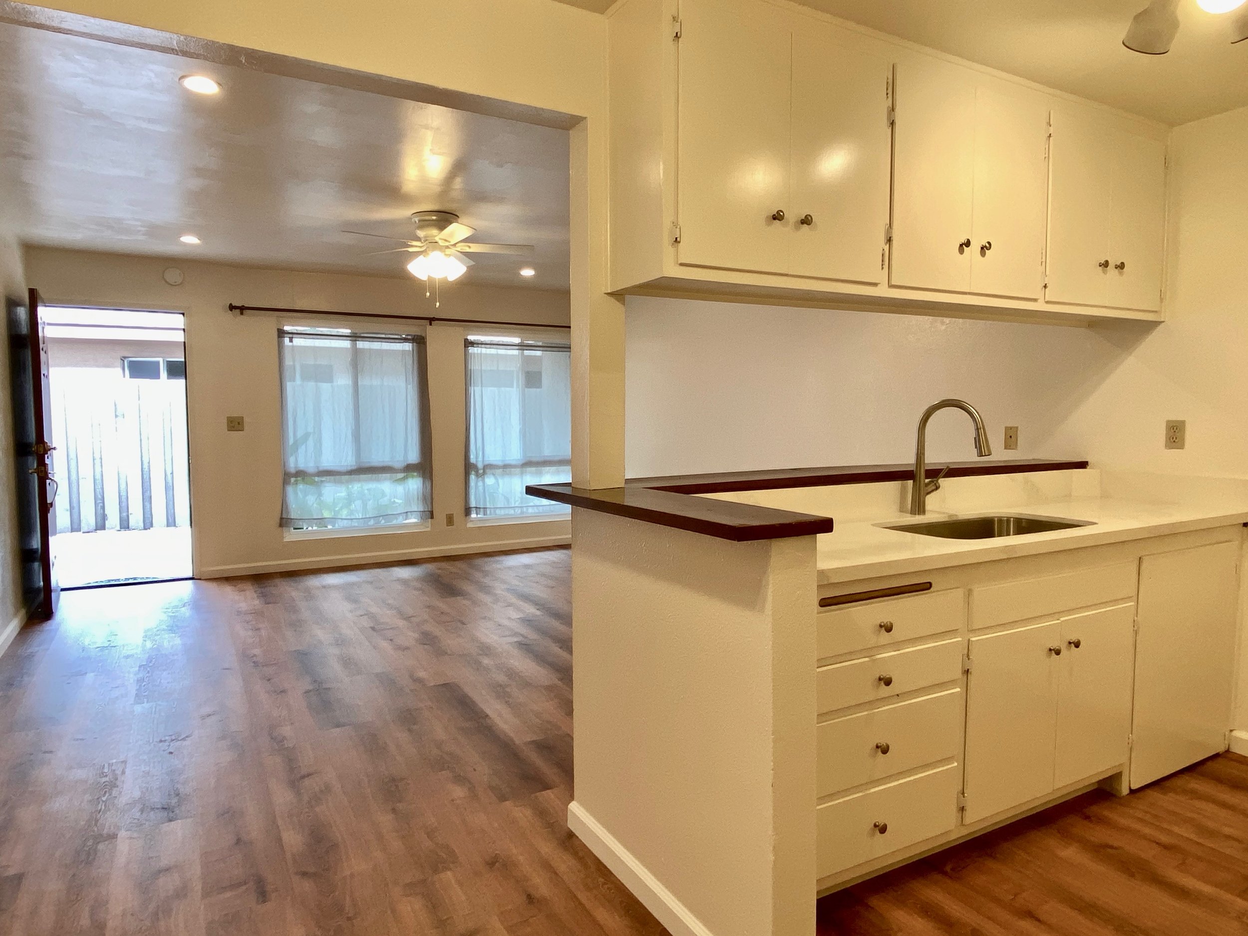 Kitchen with white cabinets, a kitchen counter with a sink, wood flooring, and an open living area with windows and a ceiling fan.